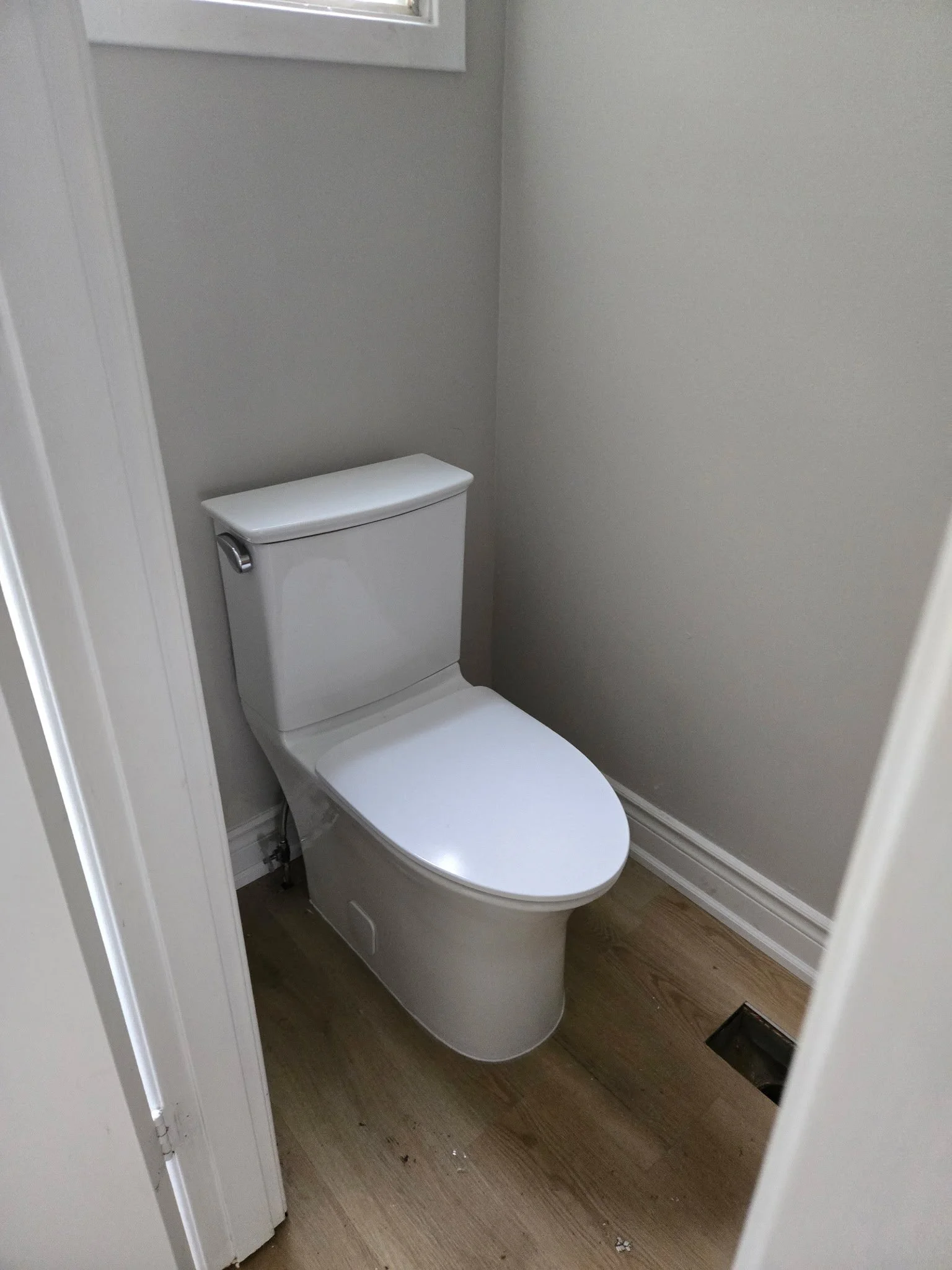 A white toilet in a small bathroom corner with beige walls and wooden floor. A small floor vent is visible near the toilet base.