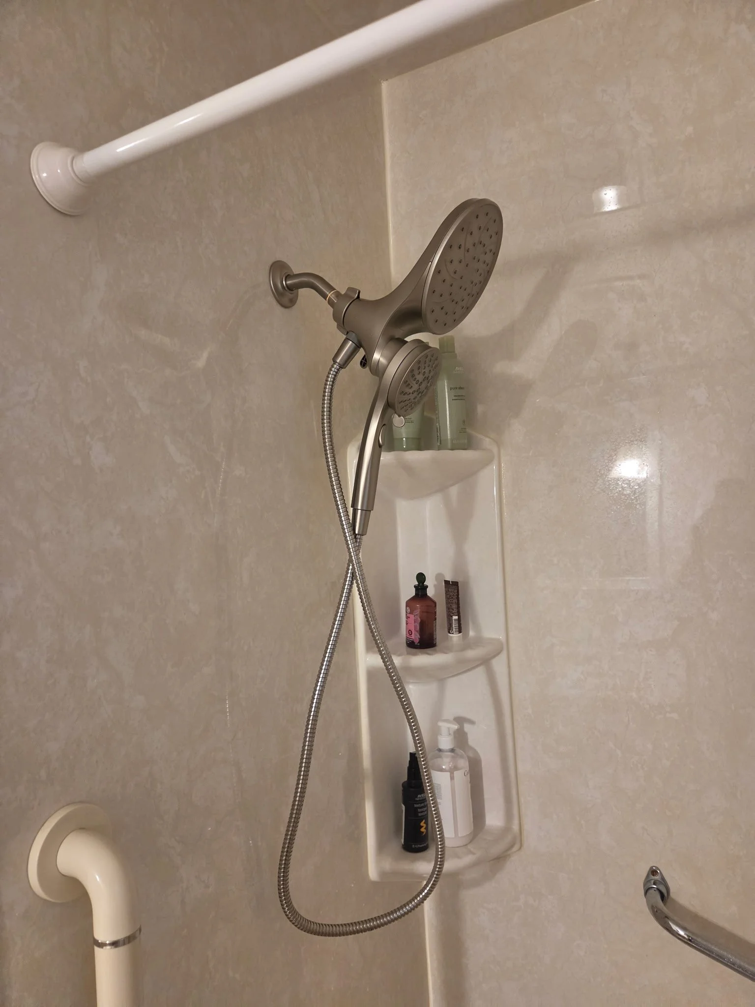 Shower area with a handheld showerhead, built-in shelf with shampoo and soap, grab bars, and a drain on the floor.