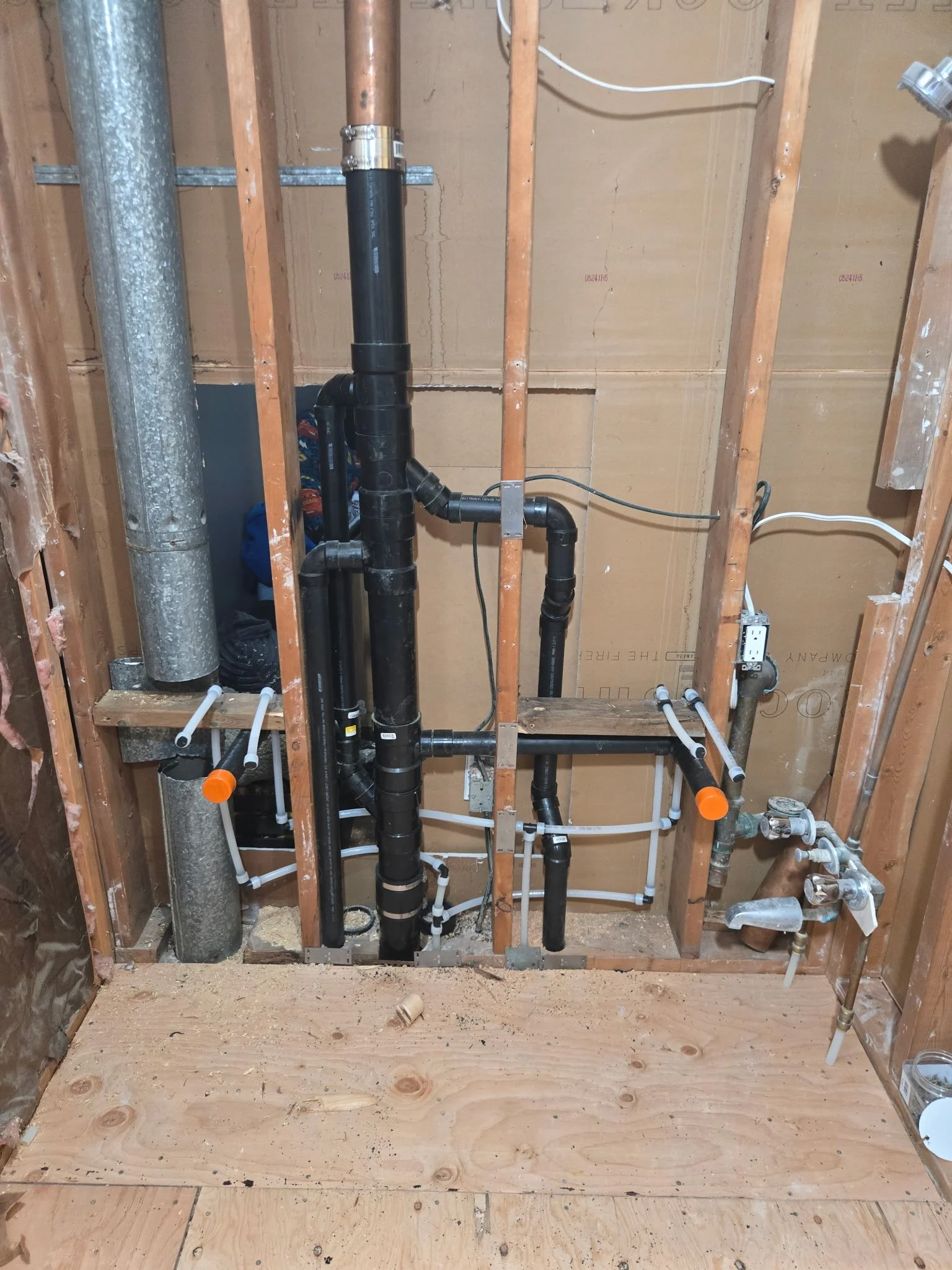 View of an unfinished bathroom wall with exposed plumbing pipes and electrical wiring, wooden studs, and a plywood wall in the background.