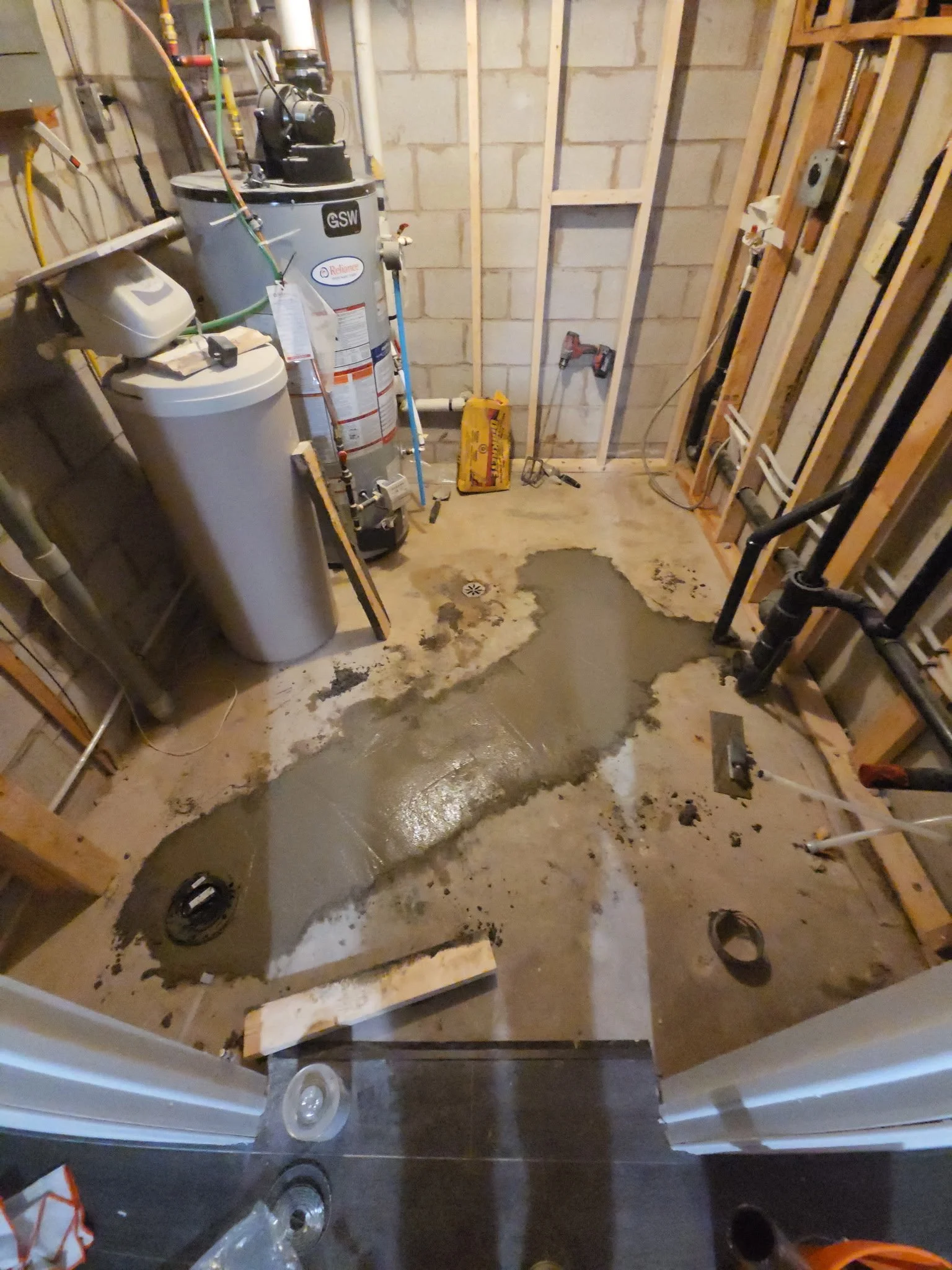 A partially constructed or renovated room with exposed framing and plumbing. The floor has concrete patches, and there are tools and materials scattered around.