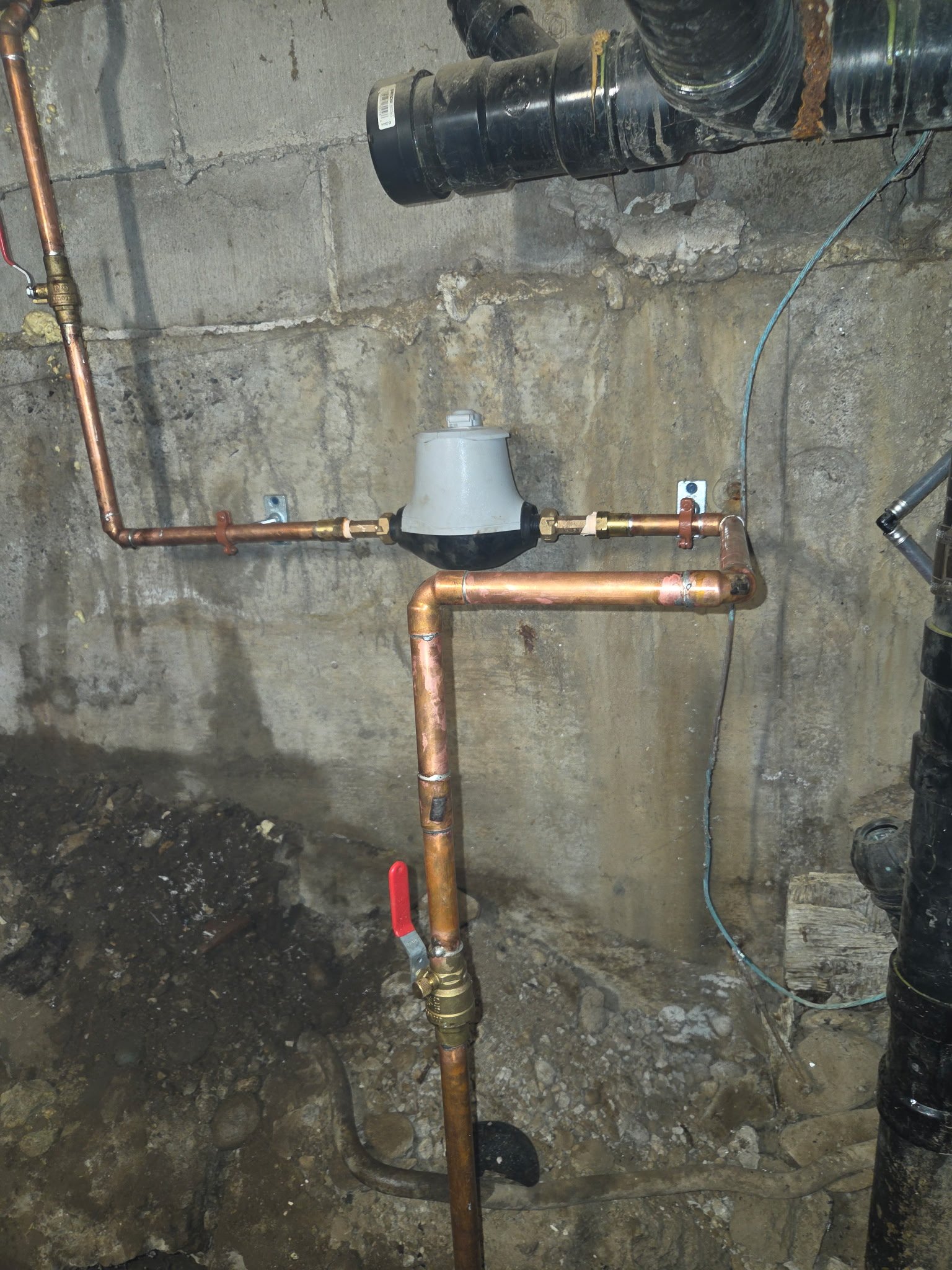 A plumbing setup with copper pipes, a white water meter, and black and copper fittings installed against a concrete wall. There are exposed pipes and wires in the background.
