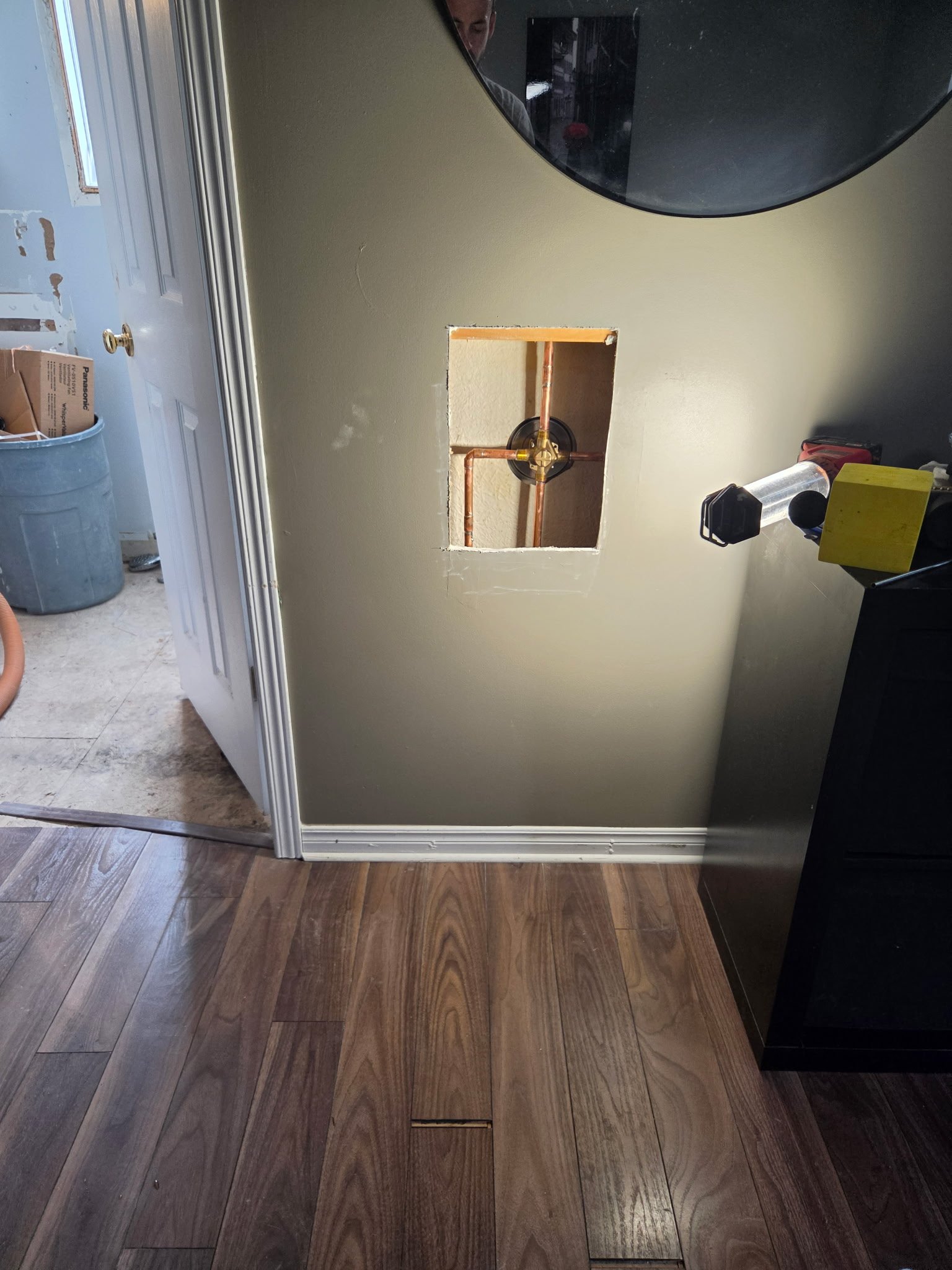 View of a bathroom wall with an opening cut out, copper pipes inside, hardwood flooring, and part of a black mirror on the right side.