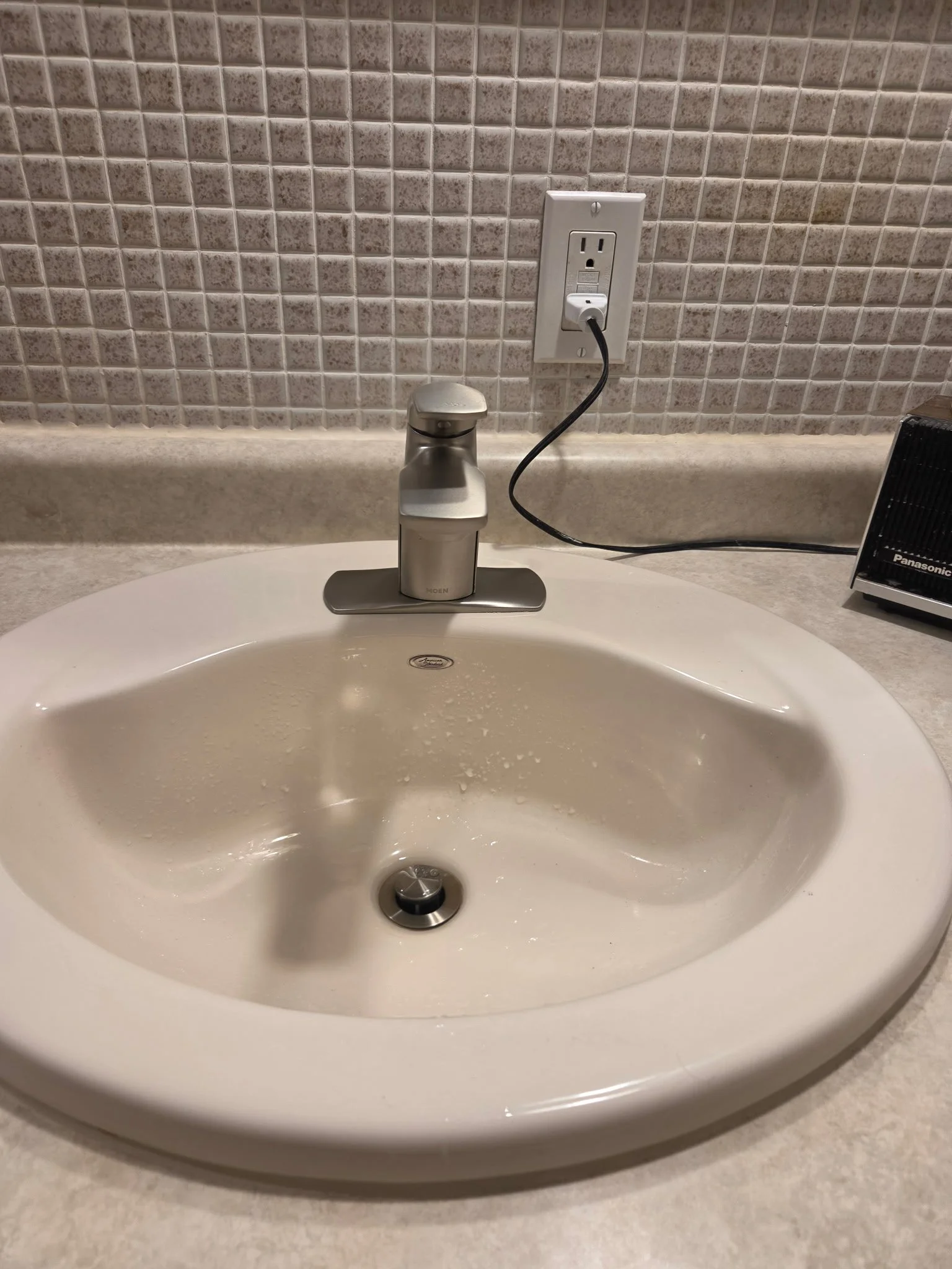 A bathroom sink with a silver faucet, a surrounding beige countertop, a tiled beige wall with a power outlet, and a black appliance on the right side.