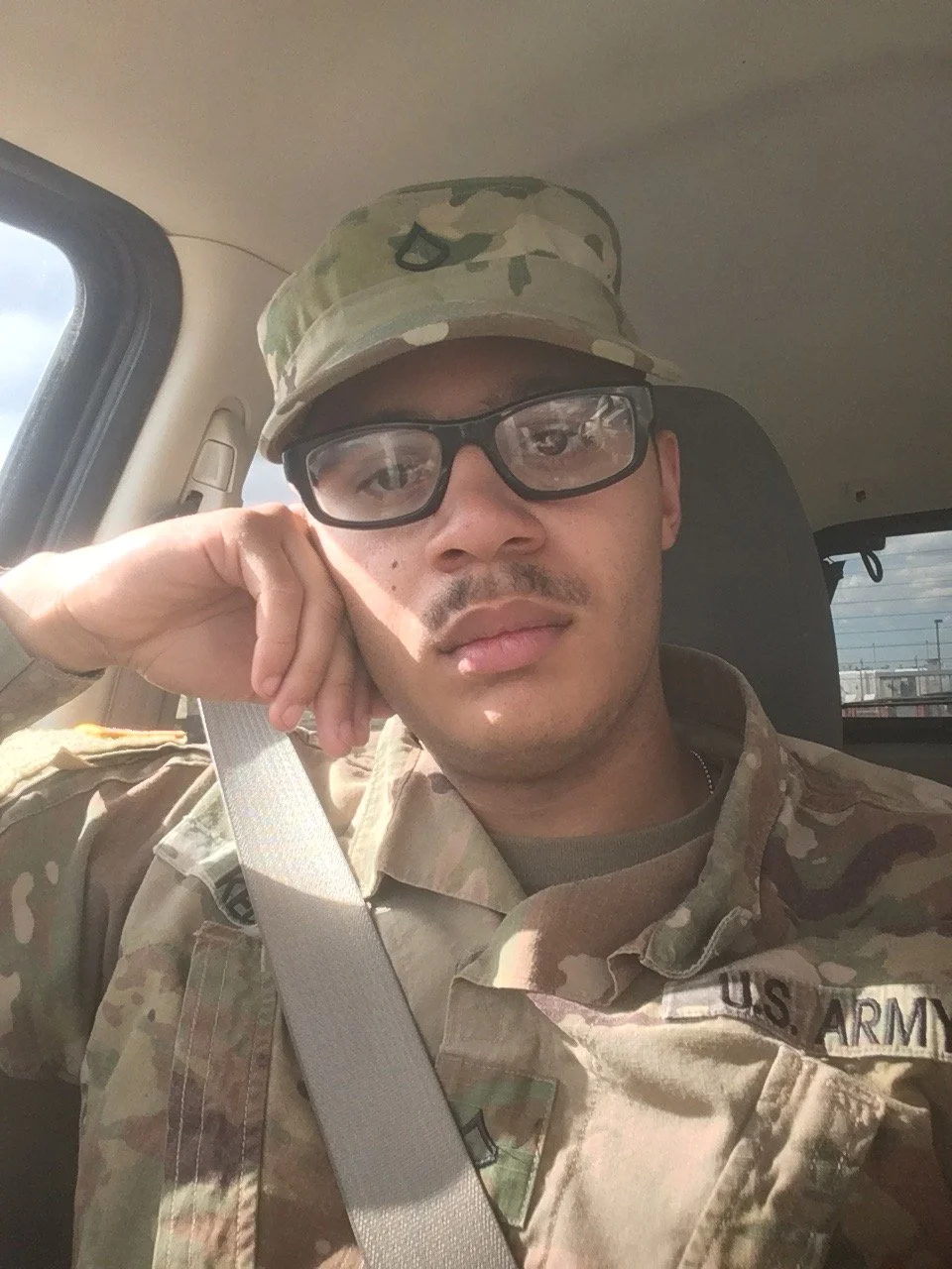 A man in U.S. Army camouflage uniform sitting in a vehicle, wearing glasses and a camouflage cap, looking at the camera with a serious expression, resting his head on his hand.