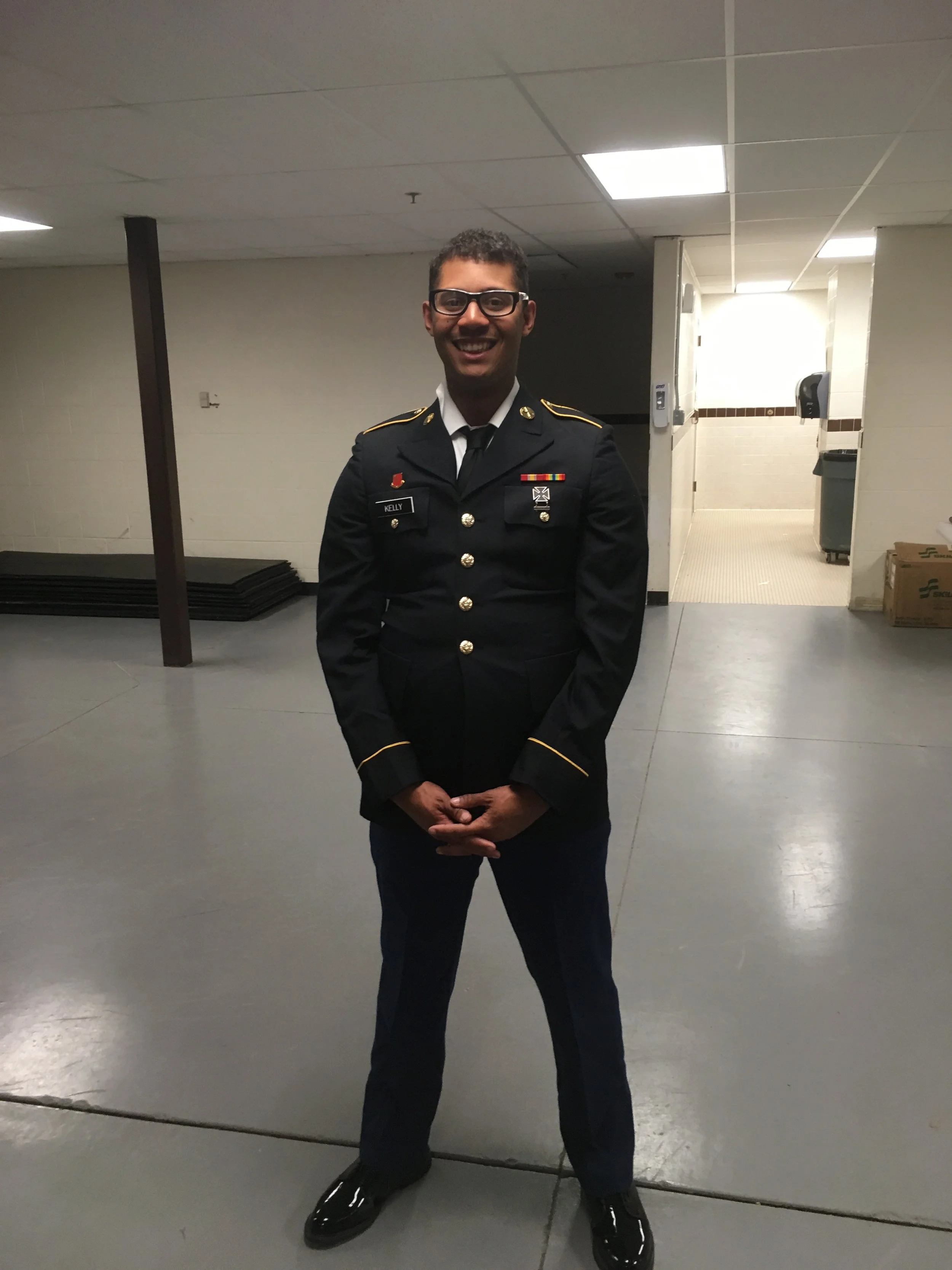 A man in a U.S. Army dress uniform, smiling, standing in a hallway with a neutral floor, beige walls, and ceiling lights.