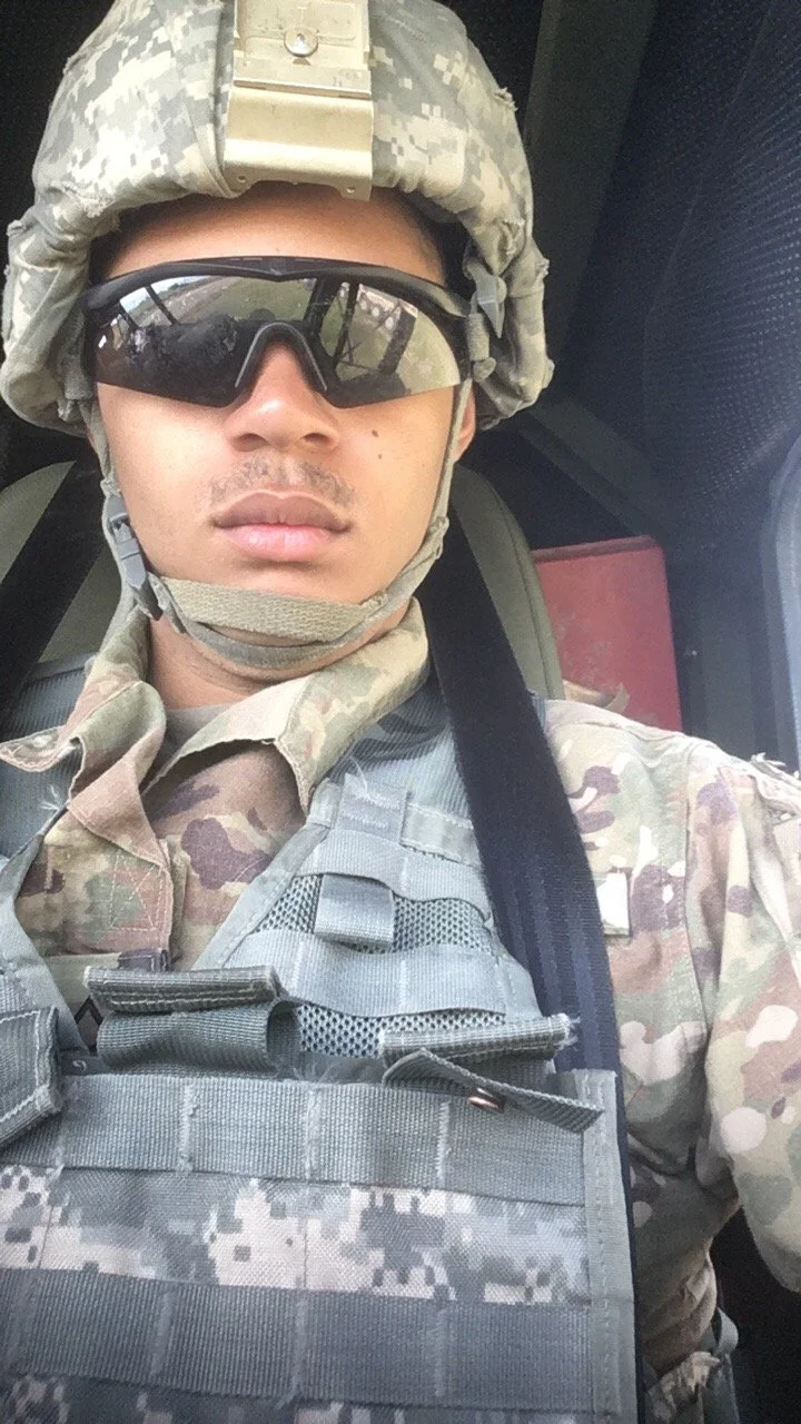 A soldier in camouflage uniform taking a selfie, wearing tactical gear, helmet, and sunglasses.