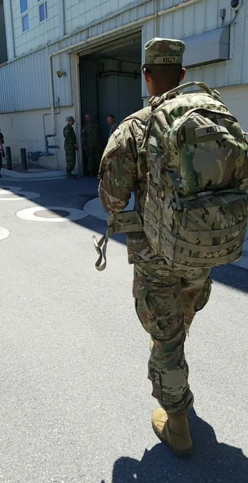 A soldier in camouflage uniform and backpack walking towards a group of soldiers standing near a building entrance.