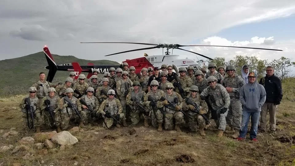 Group of soldiers in camouflage uniforms posing in front of a helicopter on a grassy hillside.