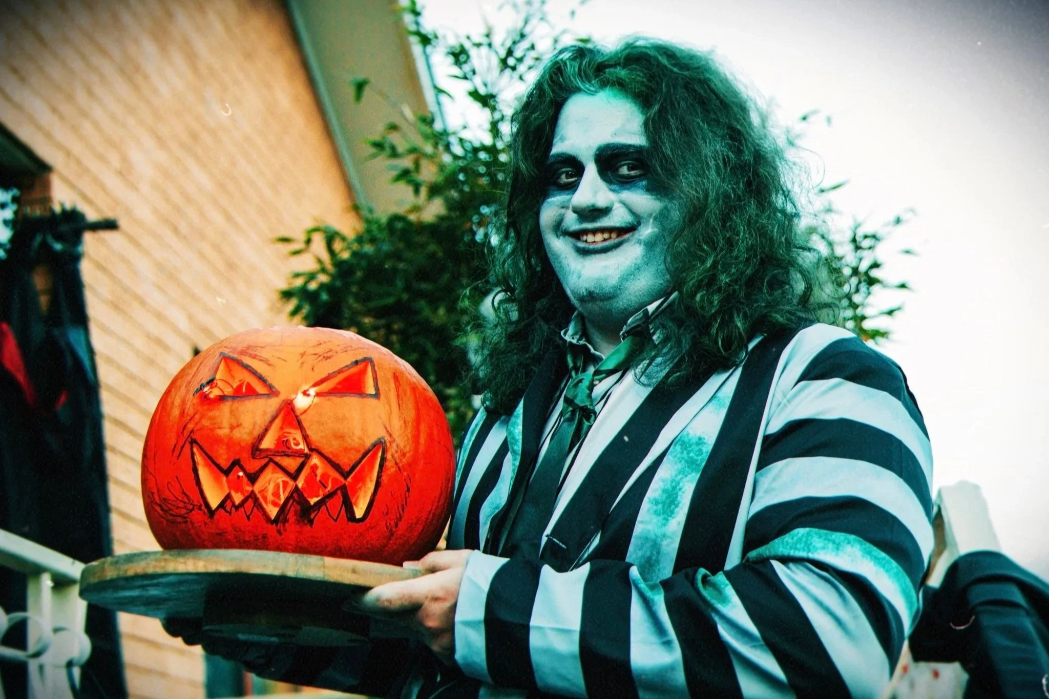 Person dressed as a goth holding a lit pumpkin with a carved face, standing outdoors with a brick wall and green plants in the background.