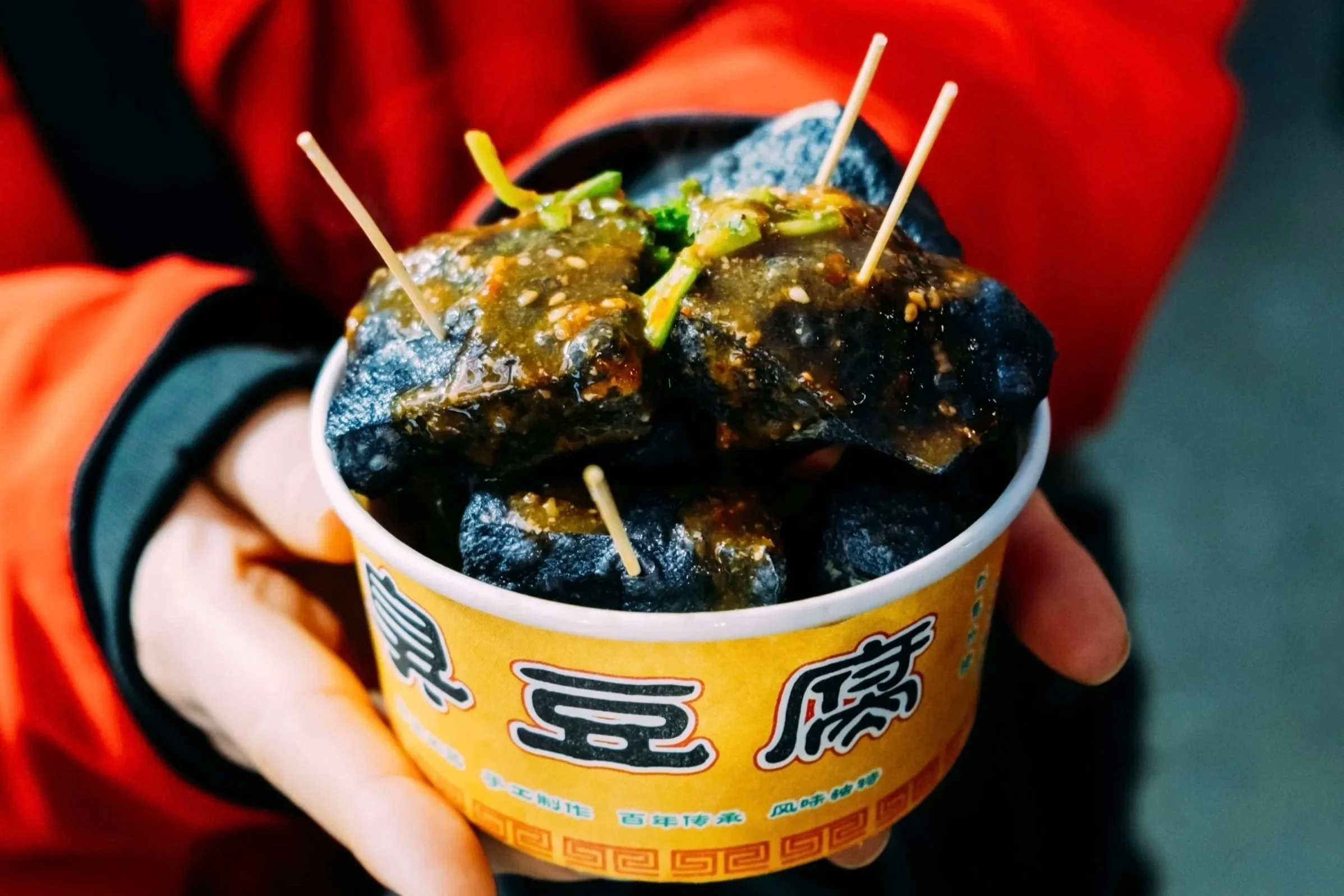 A close-up of a hand holding a cup of black jelly dessert with green toppings, served in a yellow cup with colorful Chinese characters and patterns.