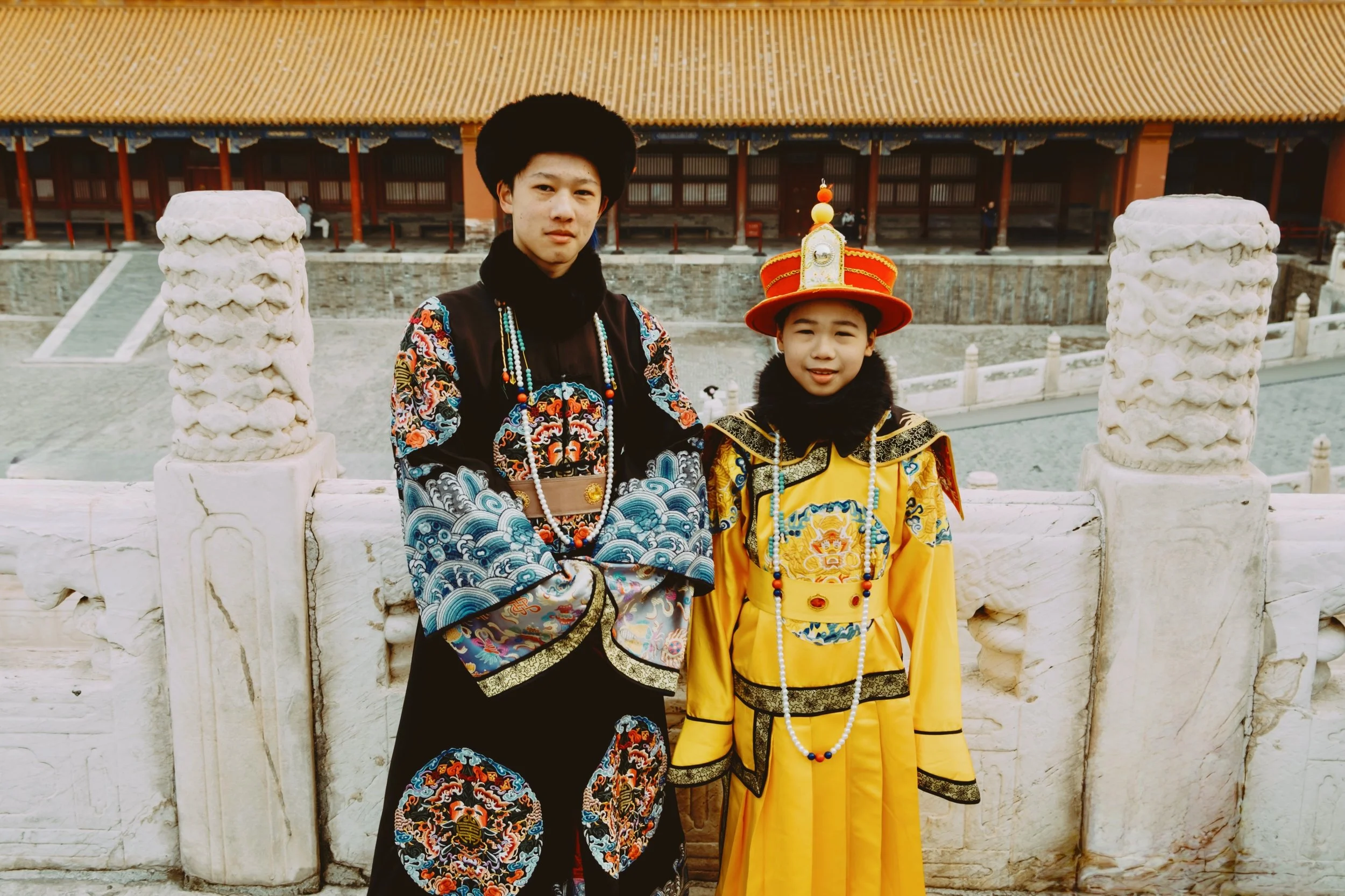 Two children dressed in traditional Chinese costumes, standing outdoors in front of a historic building with ornate pillars and a tiled roof.