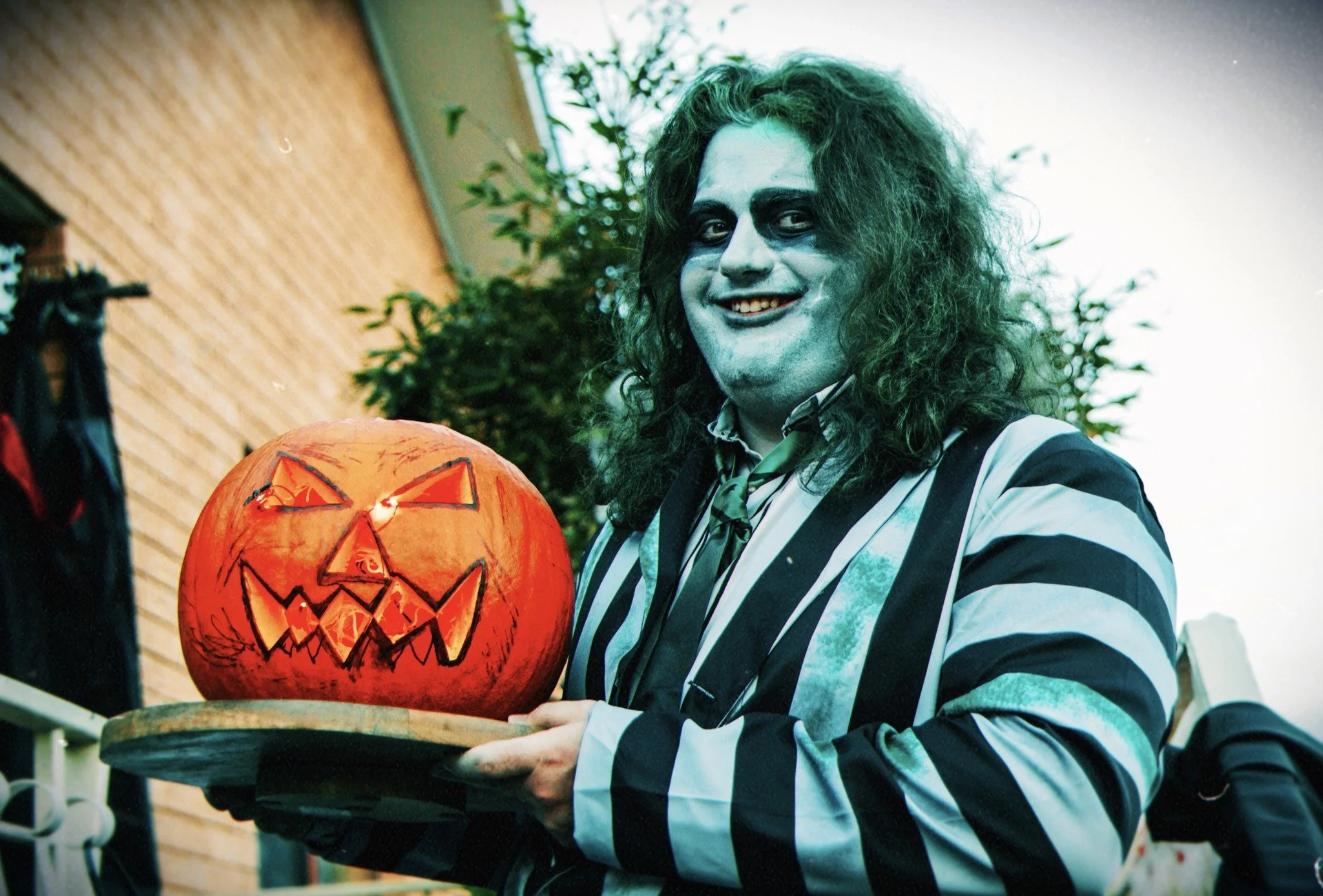 Person dressed as a spooky clown with face paint and wild hair holding a carved Halloween pumpkin lantern, outdoors during daylight.