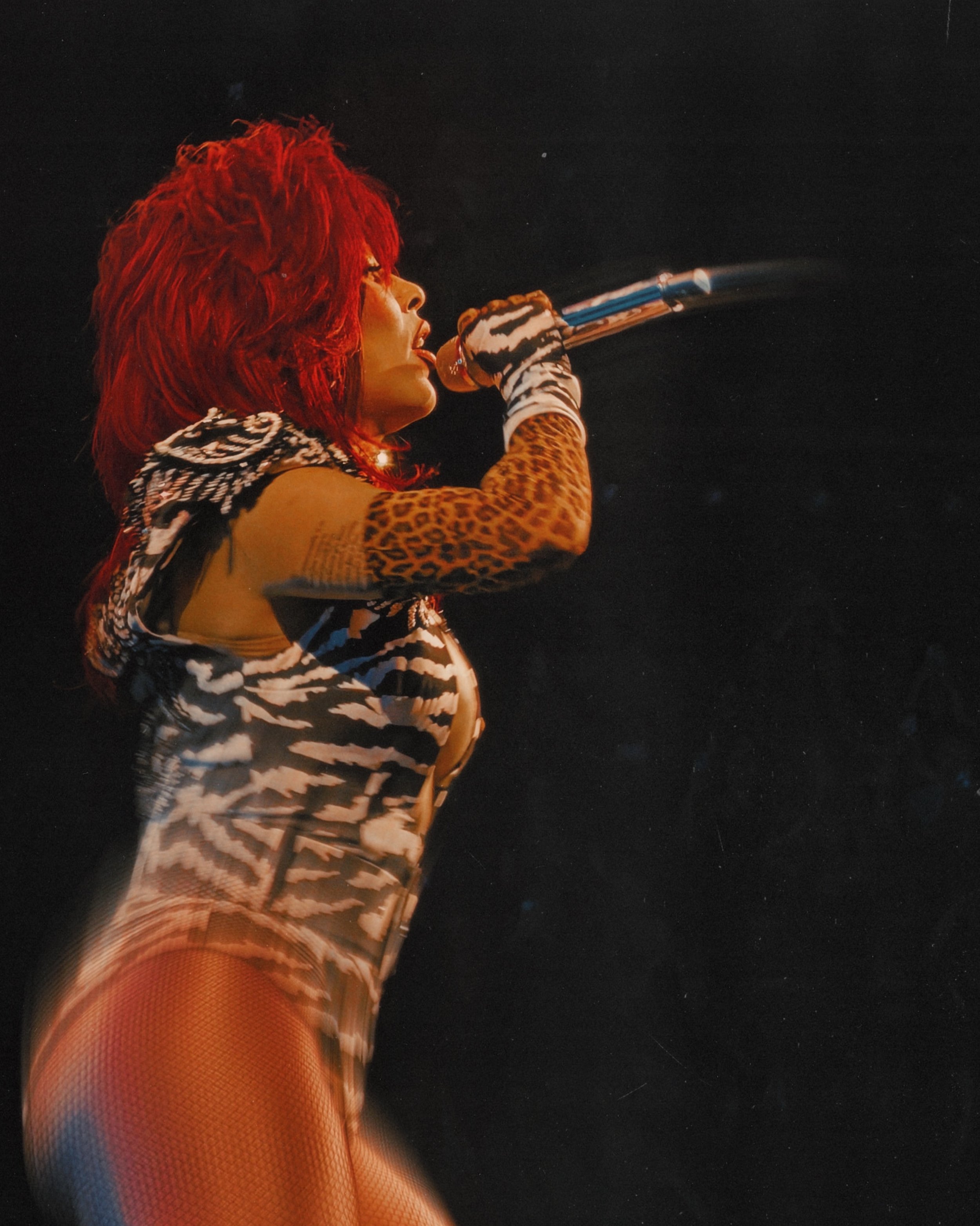 Close-up of a female performer with bright red hair singing into a microphone, wearing animal print and fishnet stockings, with a dark background.