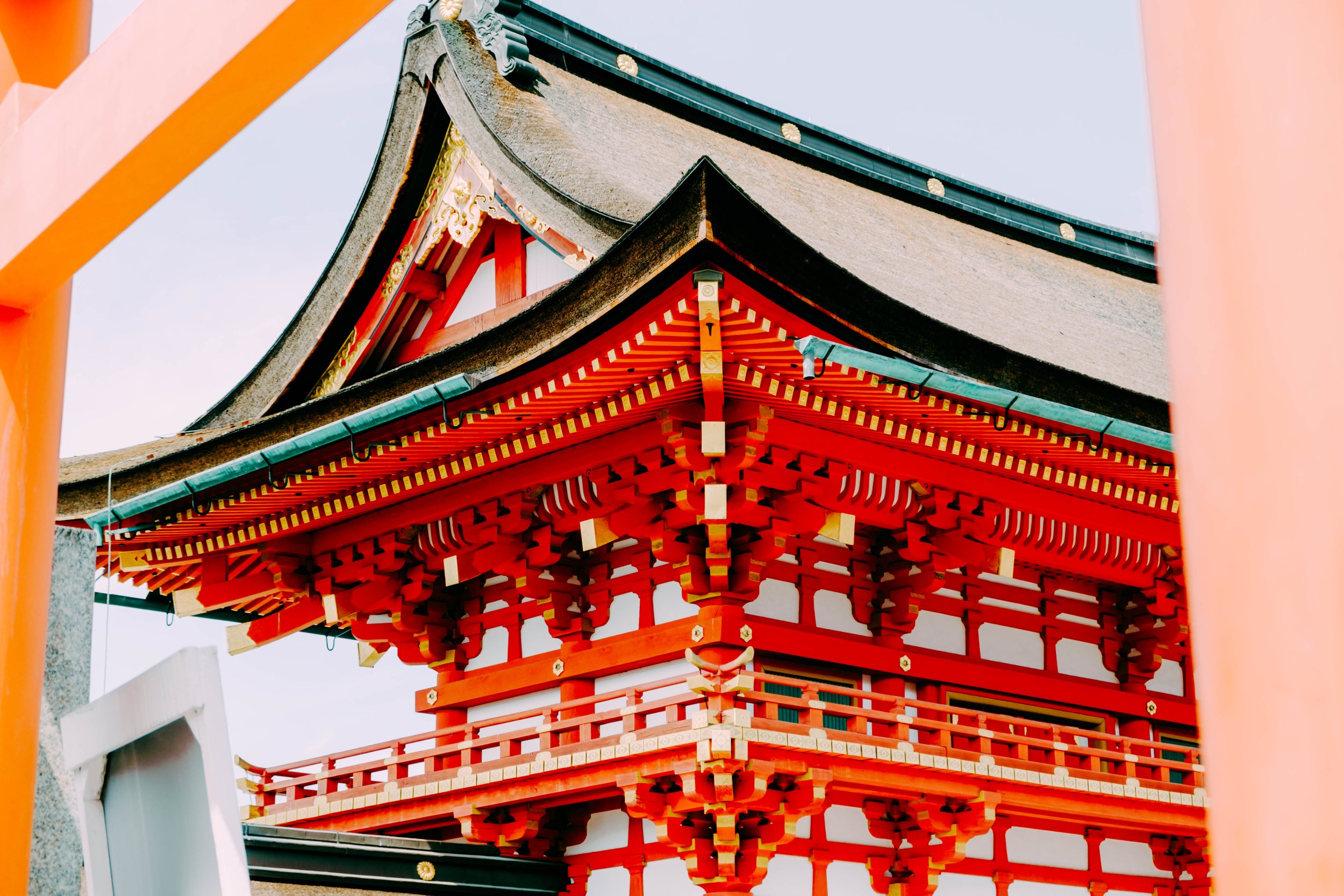 Close-up view of the roof and upper structure of a traditional Japanese shrine, showcasing its layered and ornate wooden architectural details painted in red with gold accents, against a light sky background.