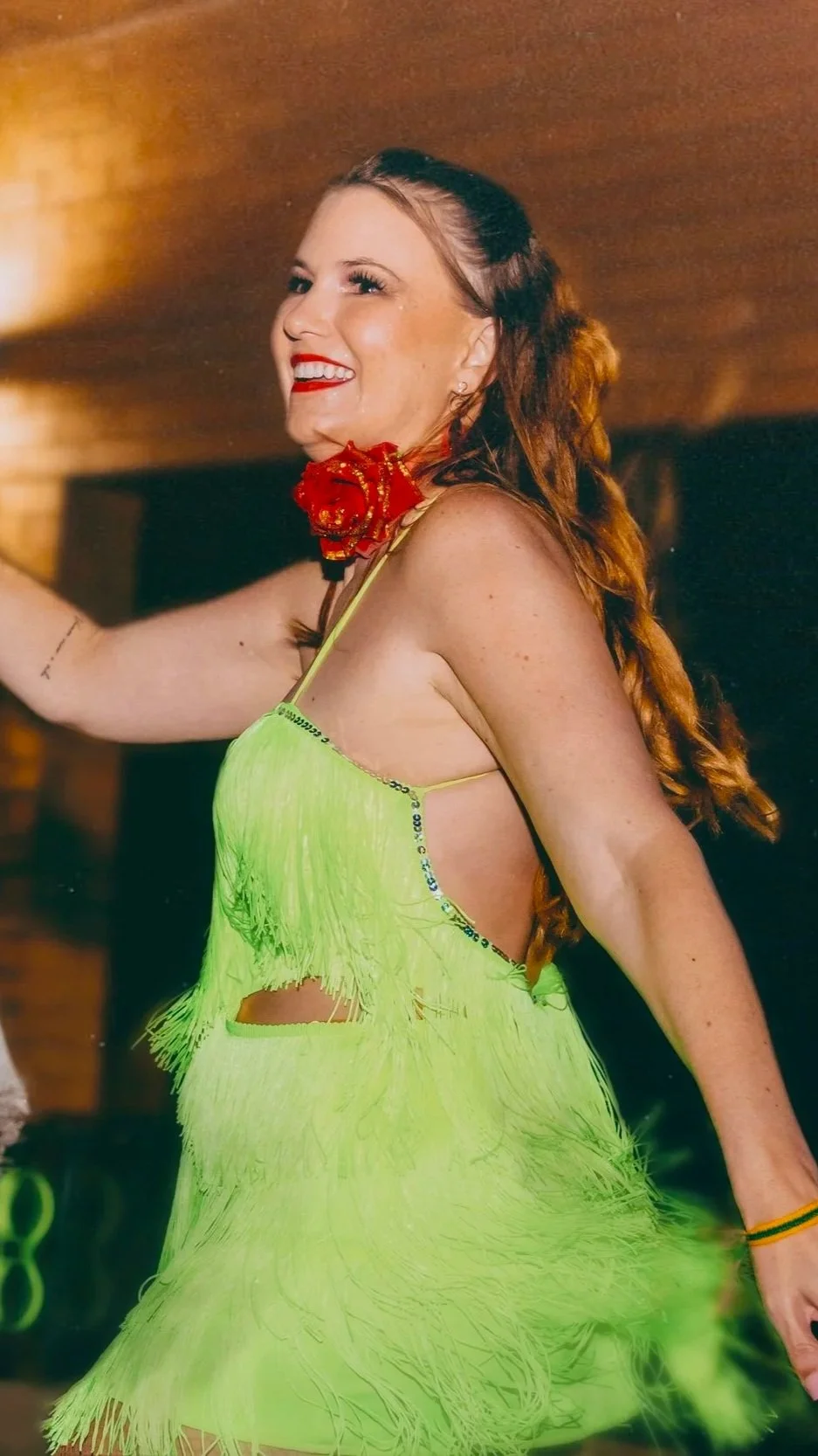 A woman with red hair styled in loose waves, smiling and wearing a bright neon green dress with fringe details and revealing sides, is holding a red rose near her neck, against a warm indoor background.