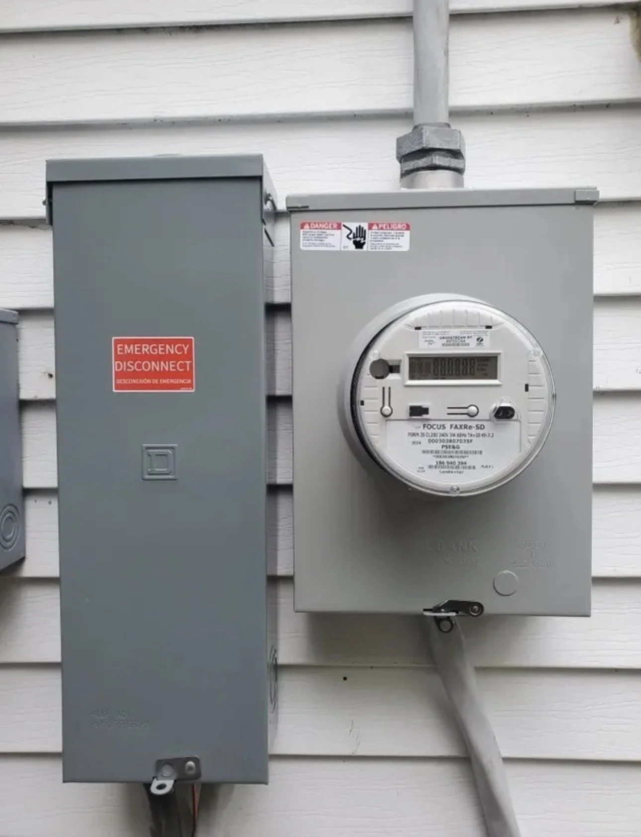 An outdoor electrical meter and circuit box mounted on a white wooden wall.