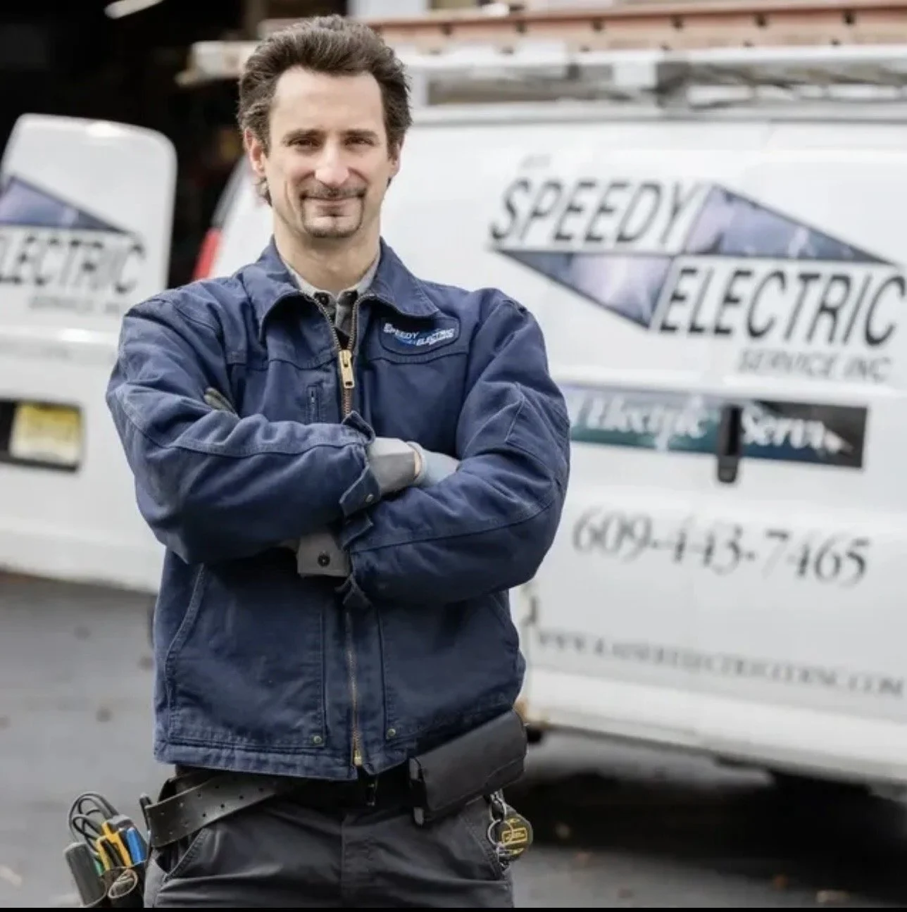 A man in a blue work jacket standing outdoors with arms crossed, in front of a white vehicle with business signage for electric services.