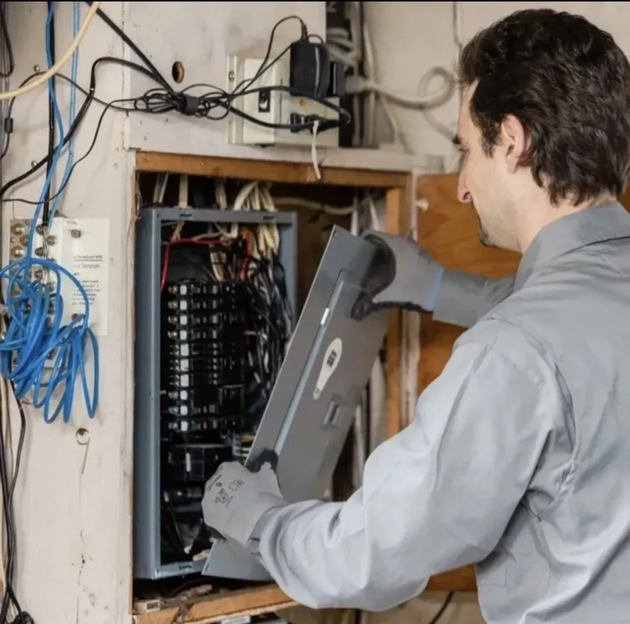 A man in protective gloves working on an electrical panel, with wires and tools around him.