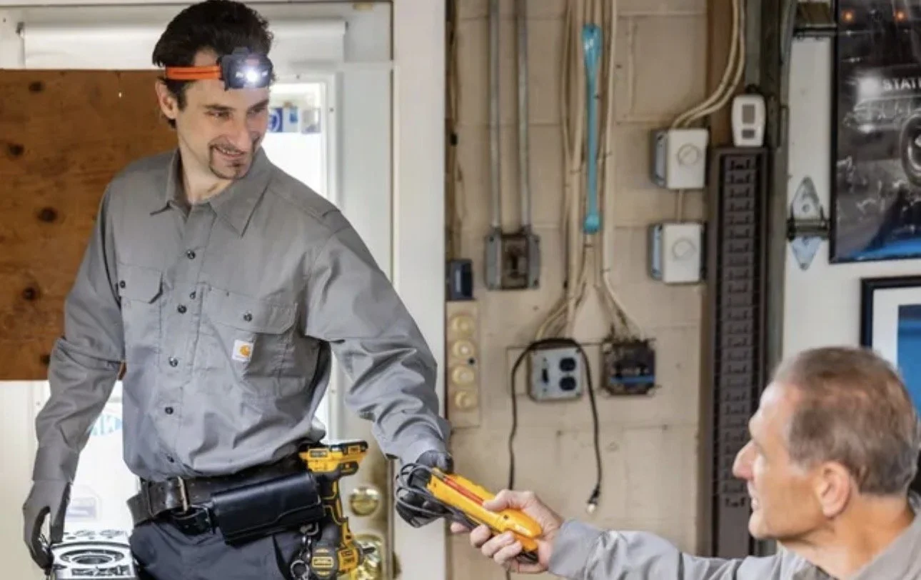 An electrician with a headlamp handing a multimeter to a client in a workshop.