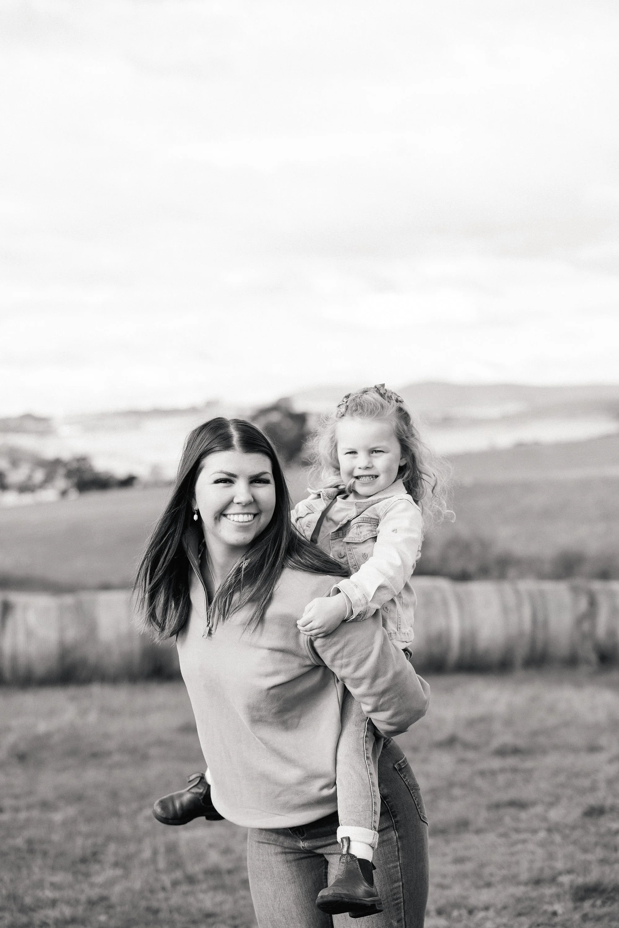 A woman holding a young girl on her shoulders outdoors in a grassy field, with a blurred landscape in the background. The image is black and white.
