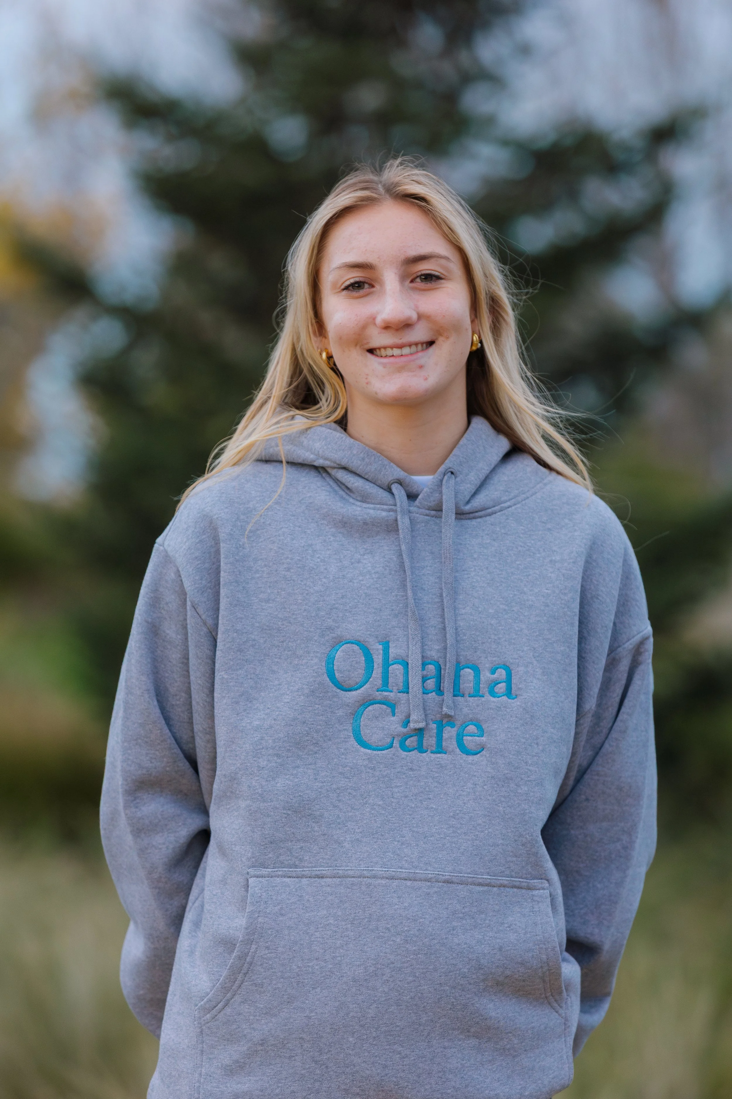 A young woman in a gray hoodie with 'Ohana Care' written on it, smiling outdoors with blurred greenery in the background.