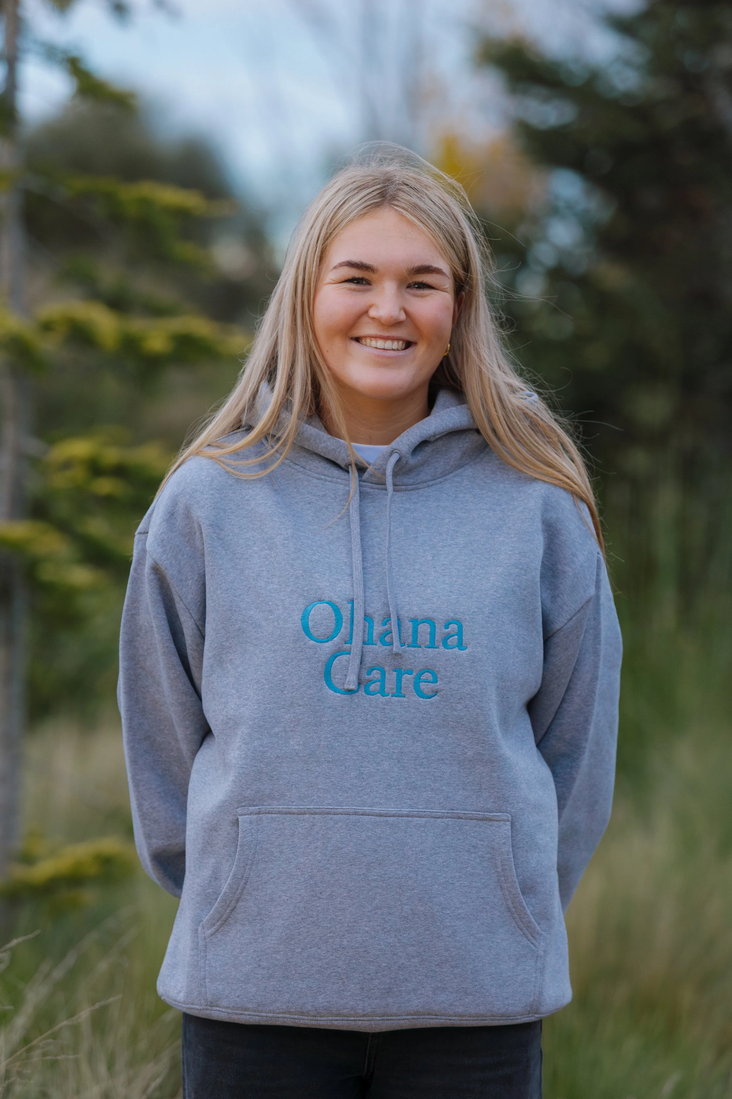 A young woman with blonde hair wearing a gray hoodie that says 'Ohlana Care' stands outdoors in a natural setting with trees and grass in the background.