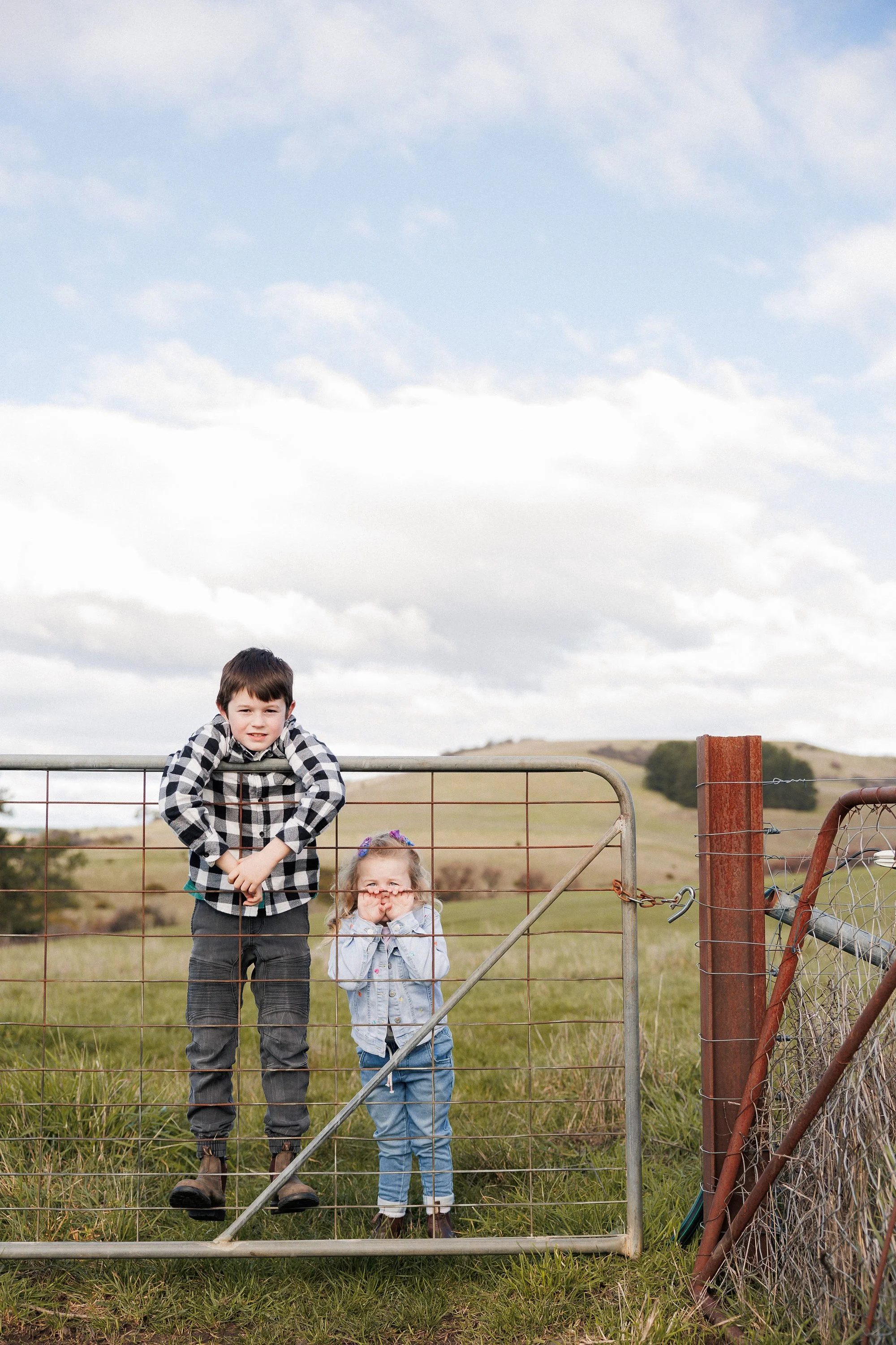 Two children standing behind a metal gate in a rural field under cloudy sky. The boy leans on the gate with hands on his cheek, and the girl stands with hands near her face, smiling.