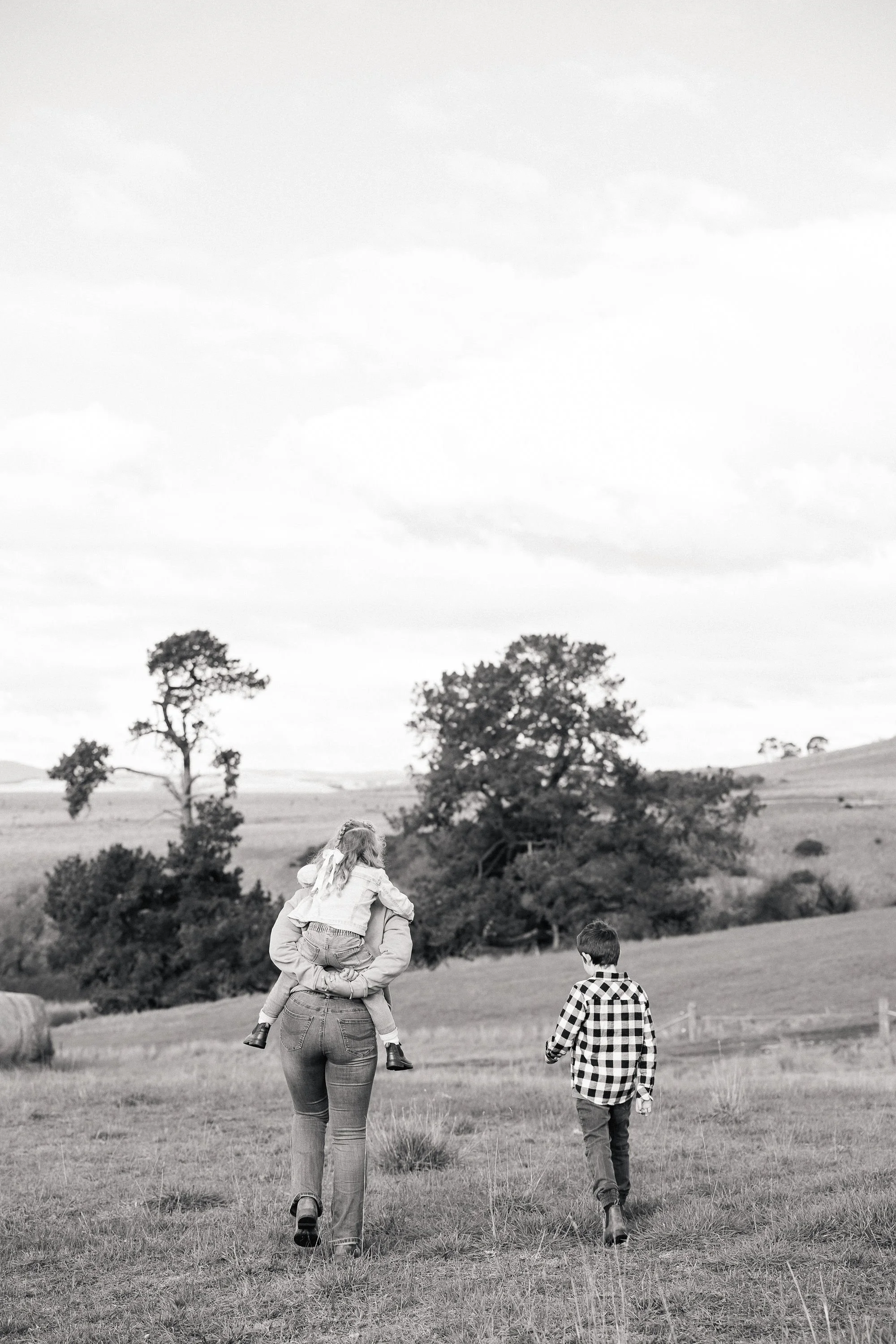 A woman giving a piggyback ride to a young girl in a grassy field with trees and rolling hills in the background, while a boy walks nearby.