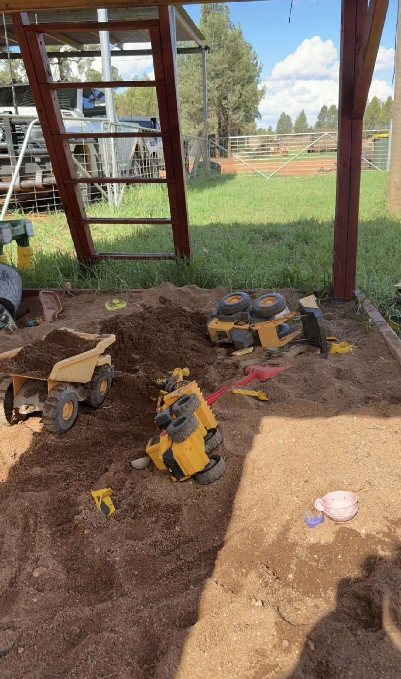 Children's sandbox area under construction with toy dump trucks, sand, and small plastic toys, with a grassy field, trees, and blue sky with clouds in the background.