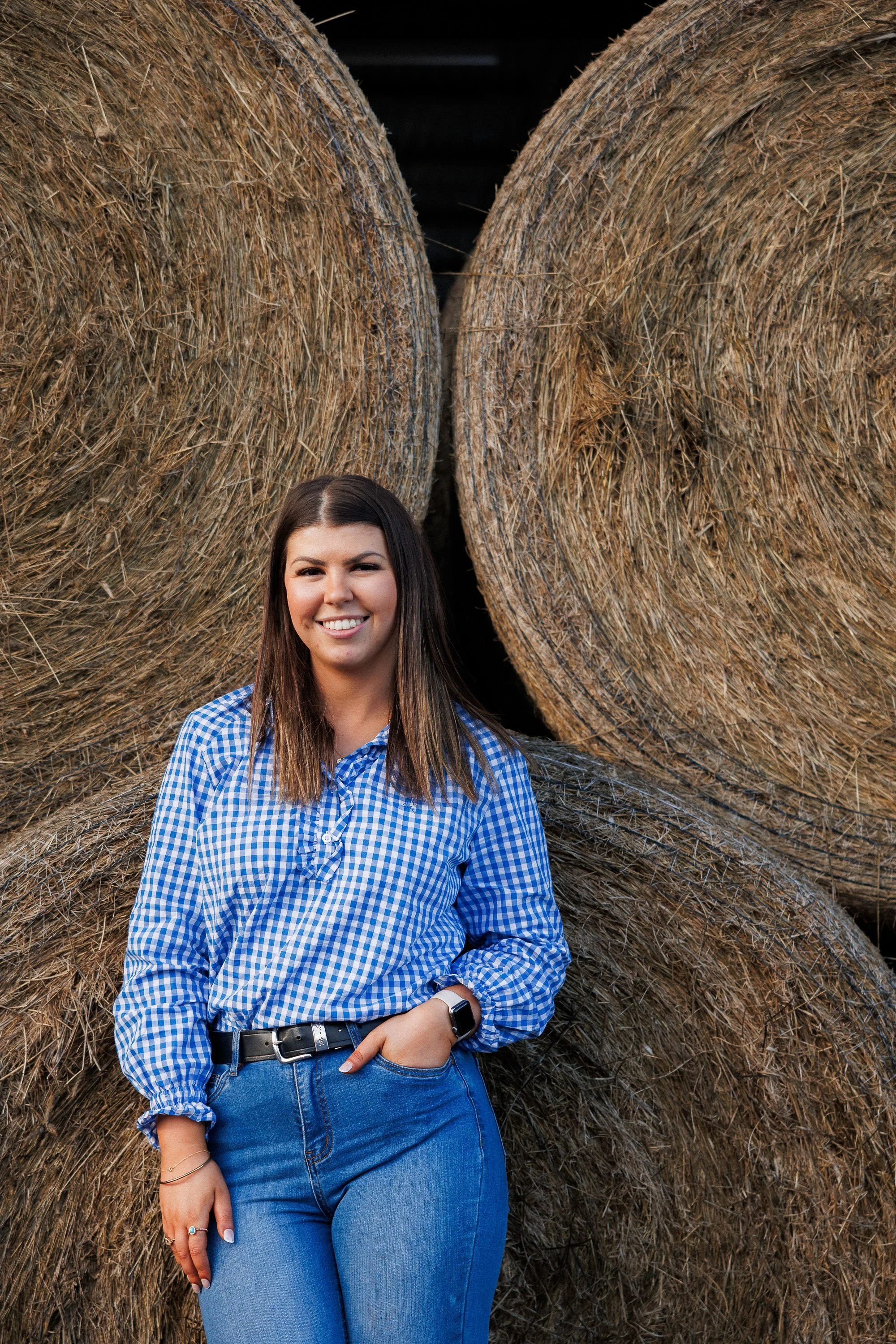 A woman in a blue and white checkered shirt and blue jeans standing in front of large round hay bales with a dark background.