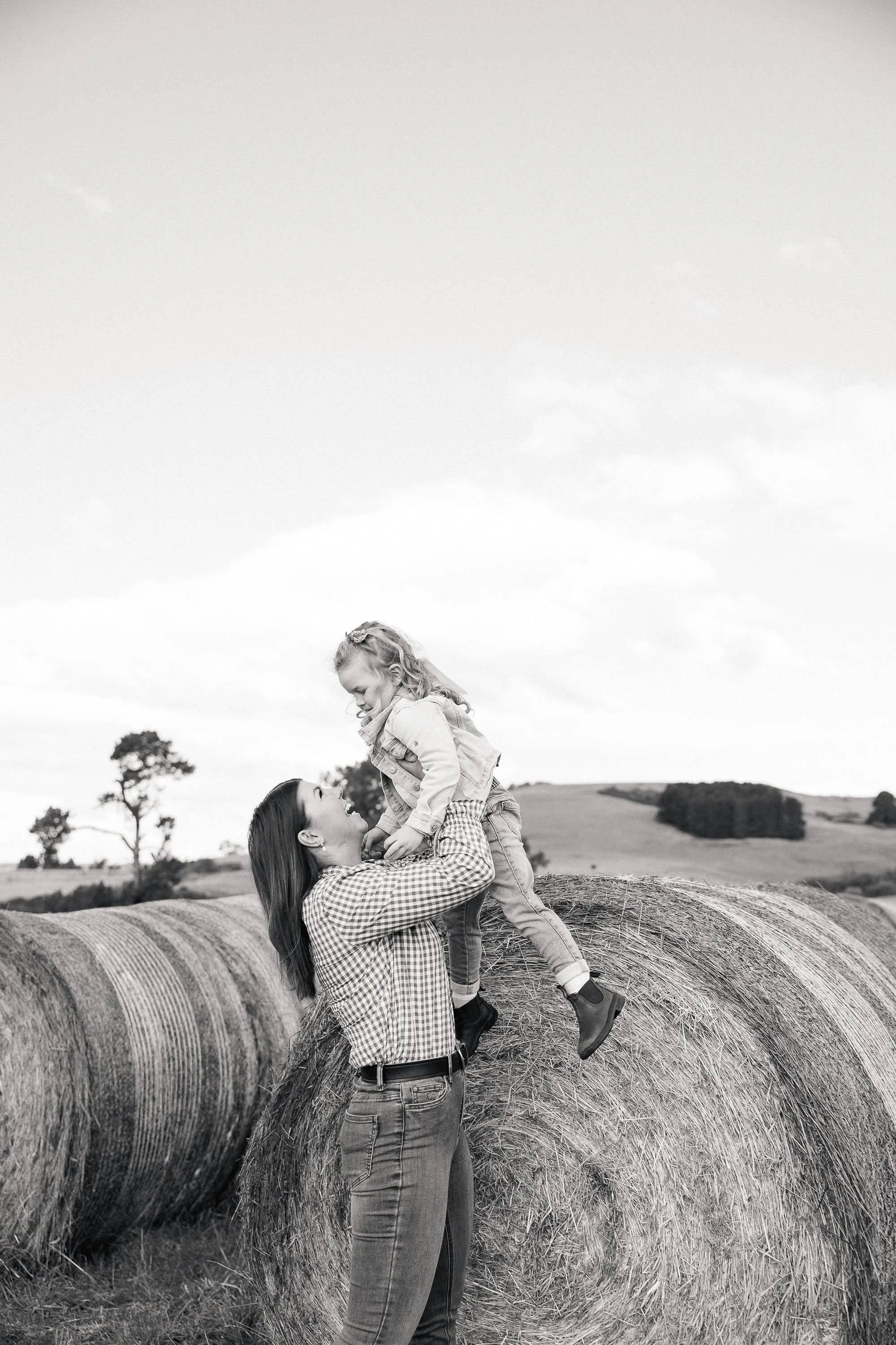 A woman with long hair and a checkered shirt lifts a girl with curly hair onto her hands on a hay bale in a field, with trees and cloudy sky in the background.