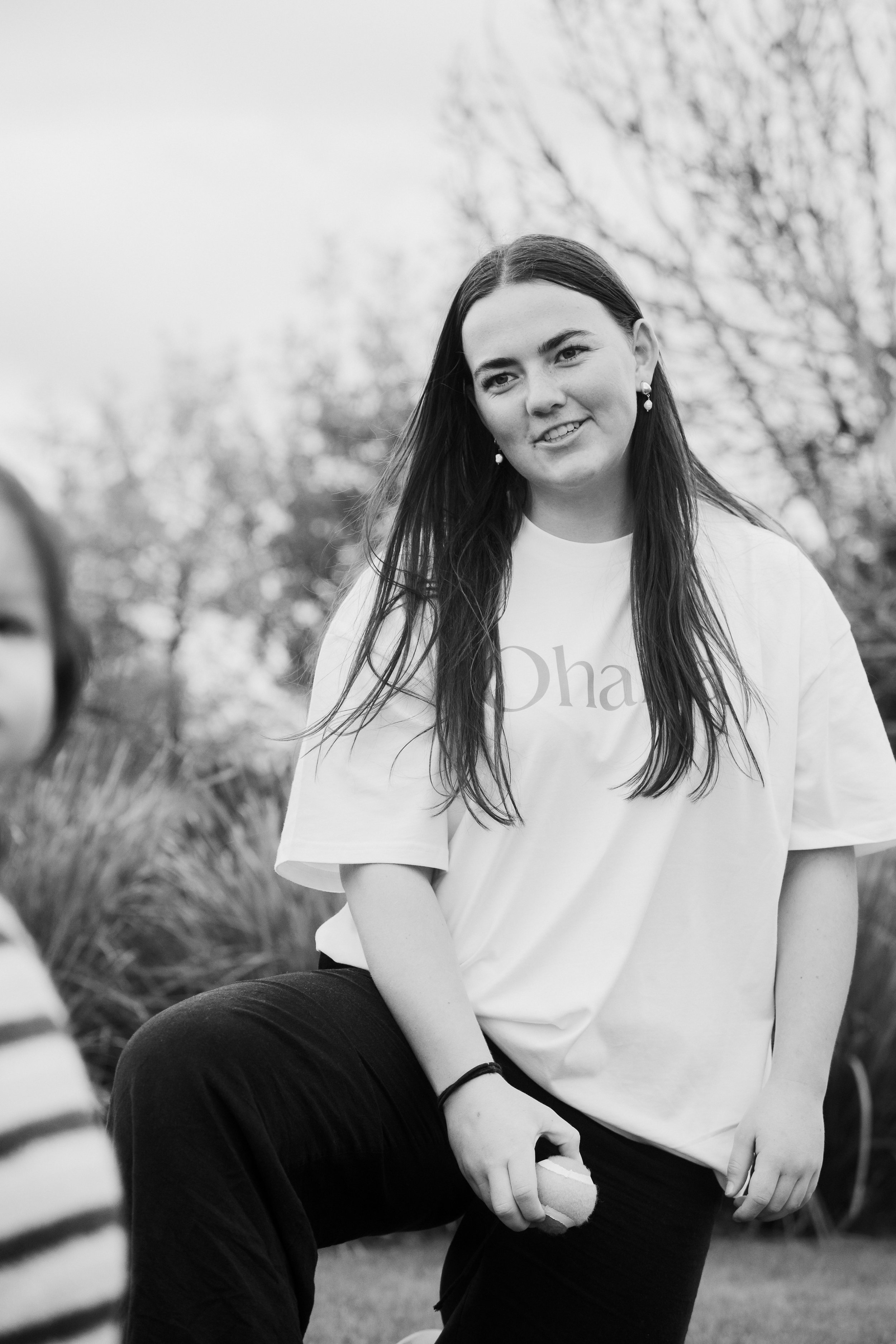 A young woman with long dark hair wearing earrings and a white T-shirt sitting outdoors among trees in black and white.