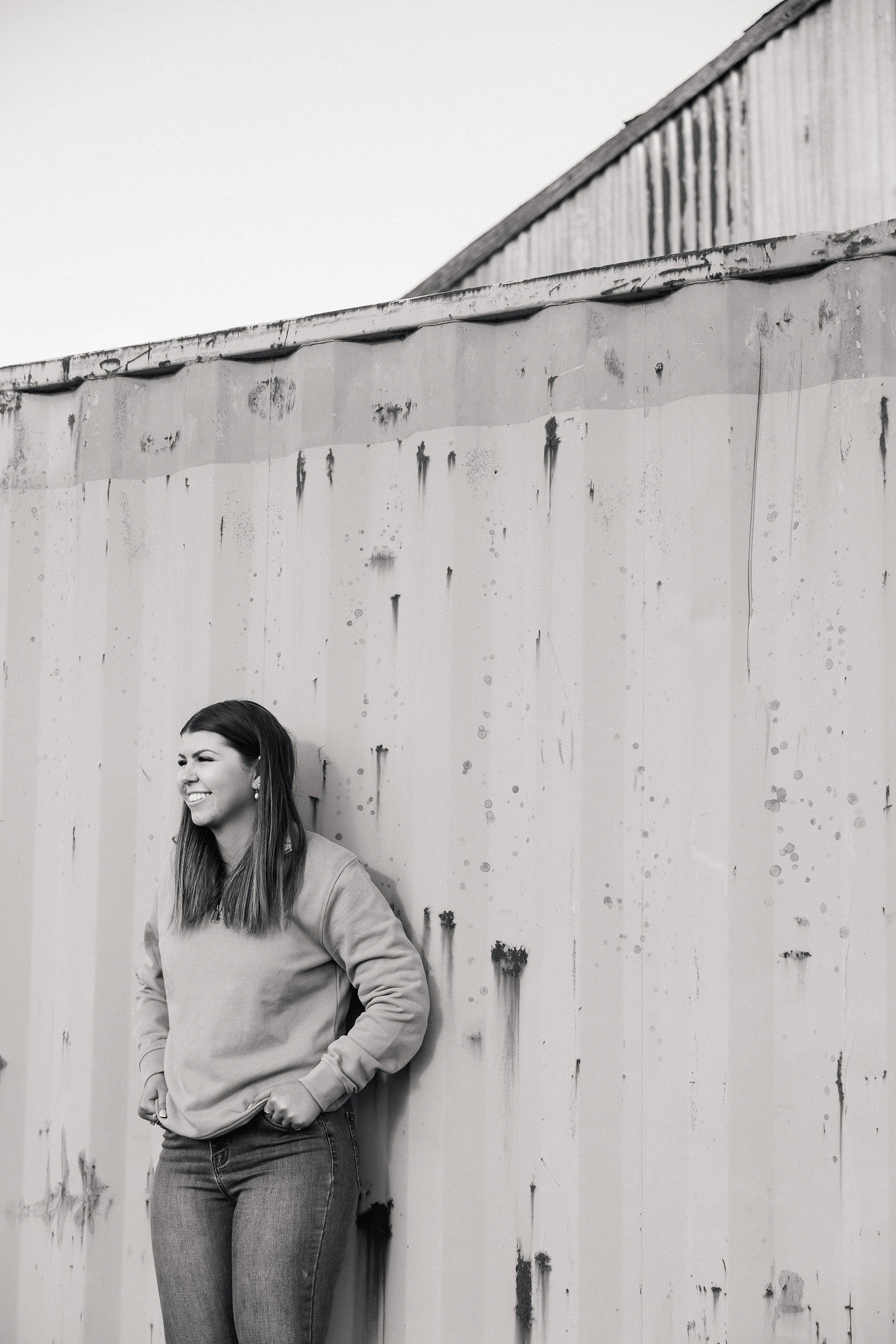 A woman with long hair wearing a jacket and jeans, standing against a weathered wall with peeling paint and stains.