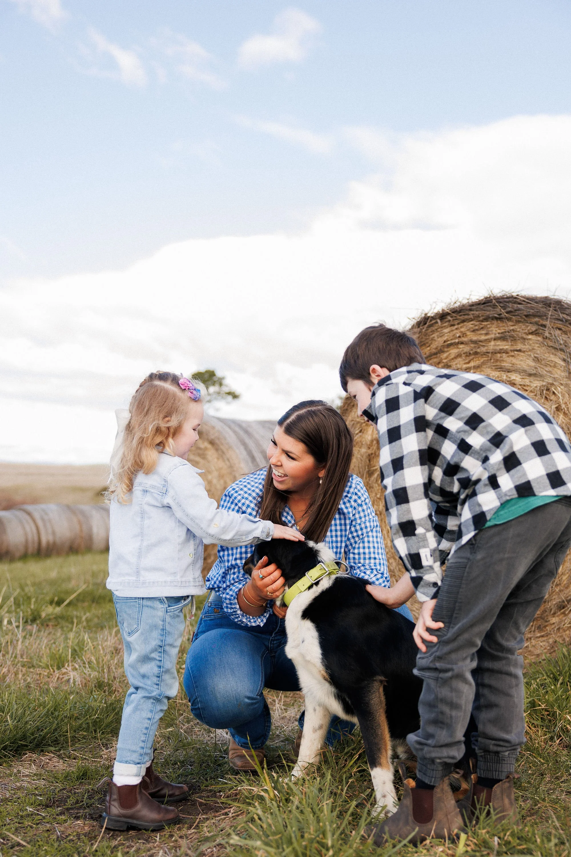 Group of children and a woman gathered outdoors in a field with haystacks in the background, interacting with a black and white puppy.
