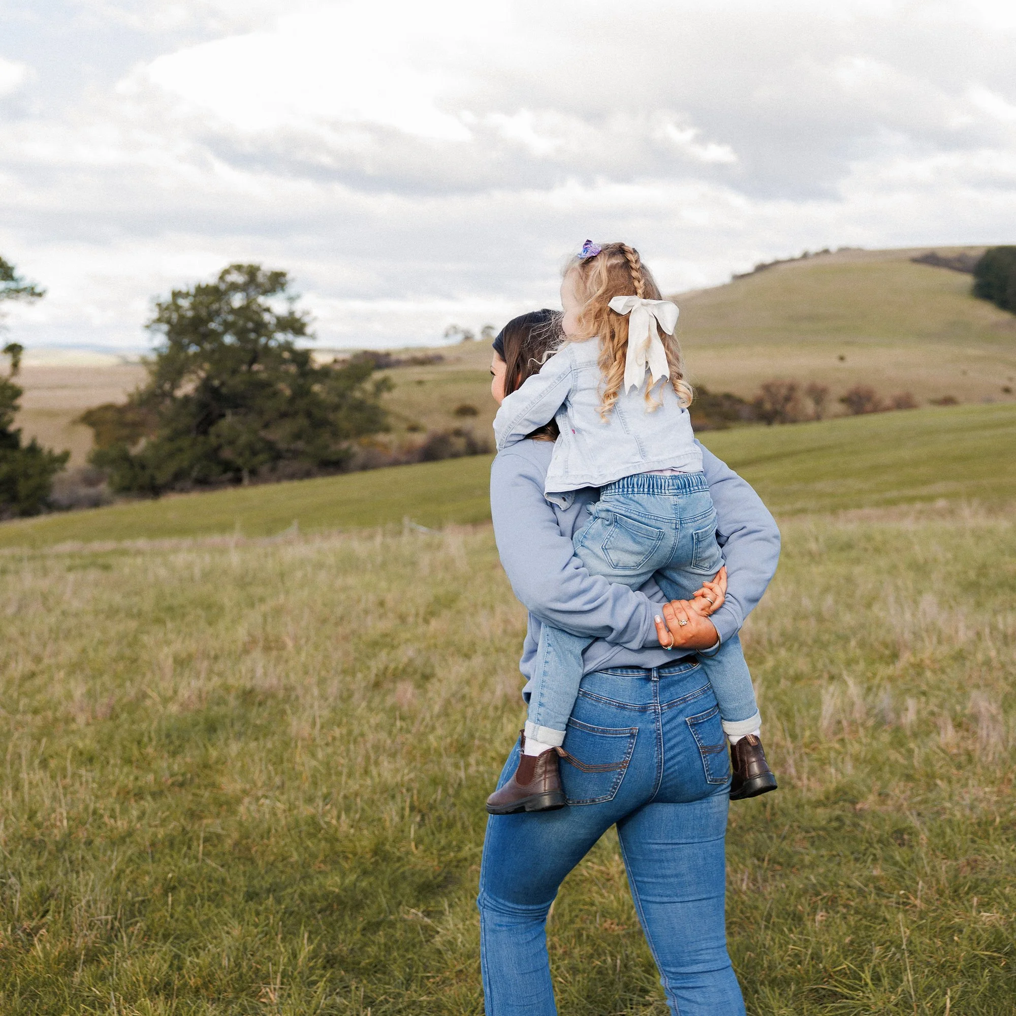 A woman with curly hair and a girl with braided hair, wearing denim jackets, are outdoors in a grassy field with trees and rolling hills under a cloudy sky, where the woman is holding the girl on her shoulders.