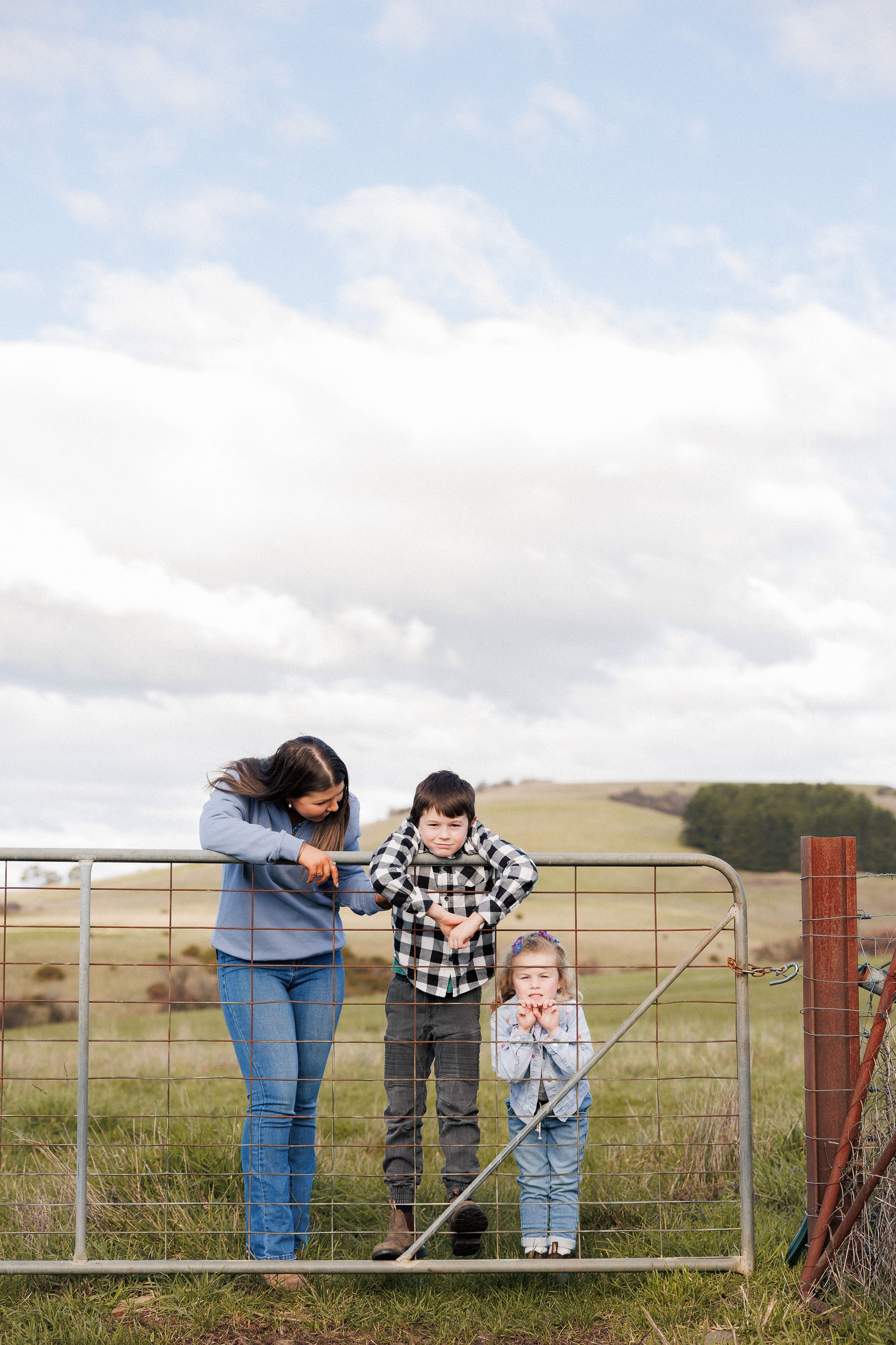 A woman and three children standing near a farm gate in a rural field under a cloudy sky.