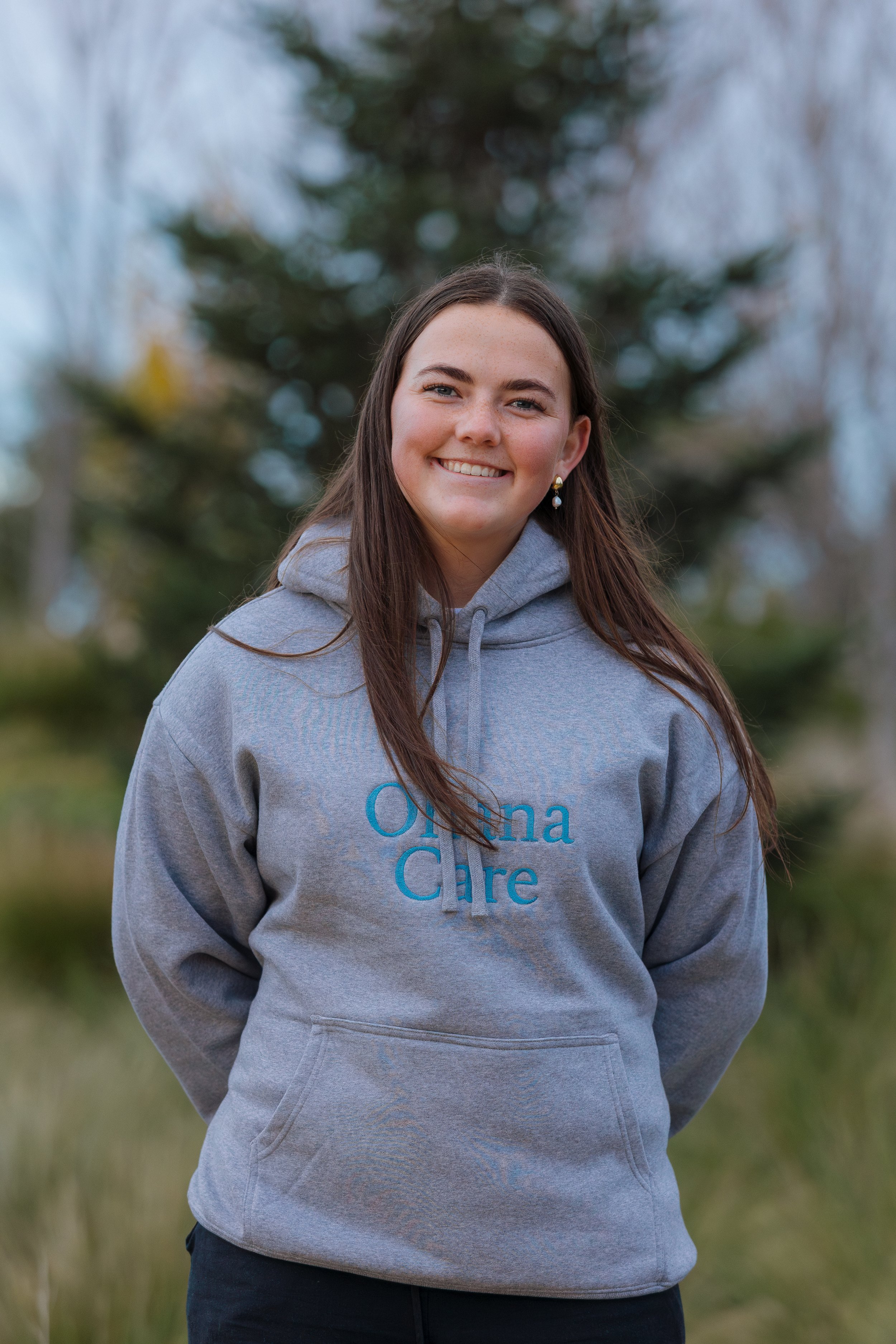 A young woman with long brown hair, smiling, wearing a gray hoodie with 'Omaha Care' printed on it, standing outdoors with blurred trees in the background.
