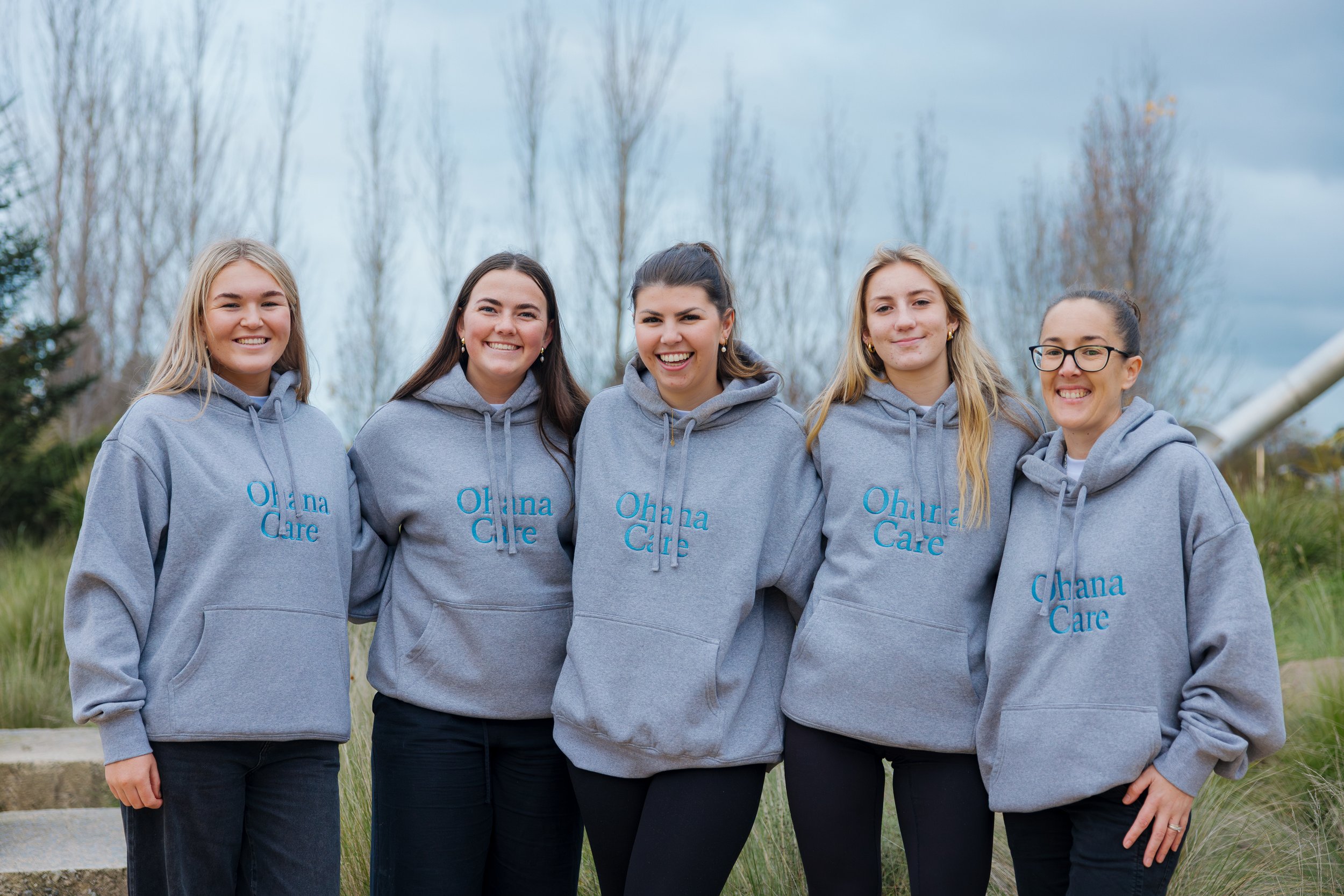 A group of five young women standing outdoors, smiling, wearing matching gray hoodies with blue text that reads 'Ohana Care,' with trees and cloudy sky in the background.