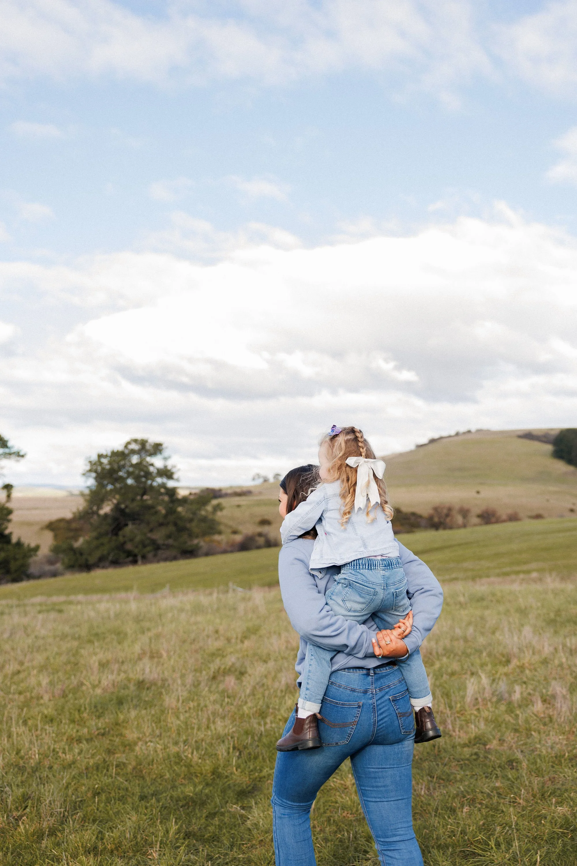 A woman with blue jeans and a denim jacket is holding a young girl on her shoulders in an open grassy field. The girl has curly blond hair tied with ribbons and is wearing a white shirt and blue jeans. The sky is partly cloudy with rolling hills and trees in the background.