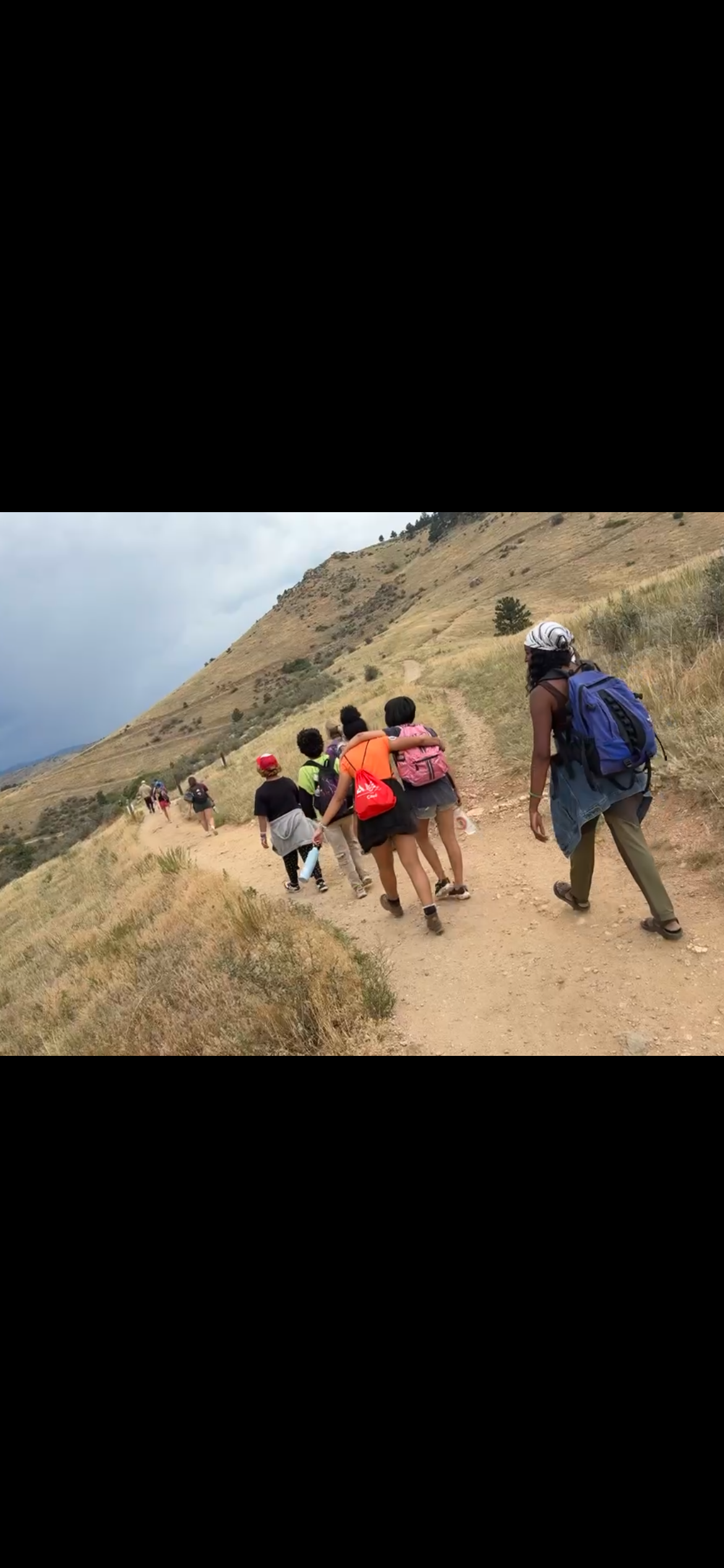 A group of diverse people hiking on a trail through a hilly landscape with dry grass, some carrying backpacks and wearing outdoor gear.