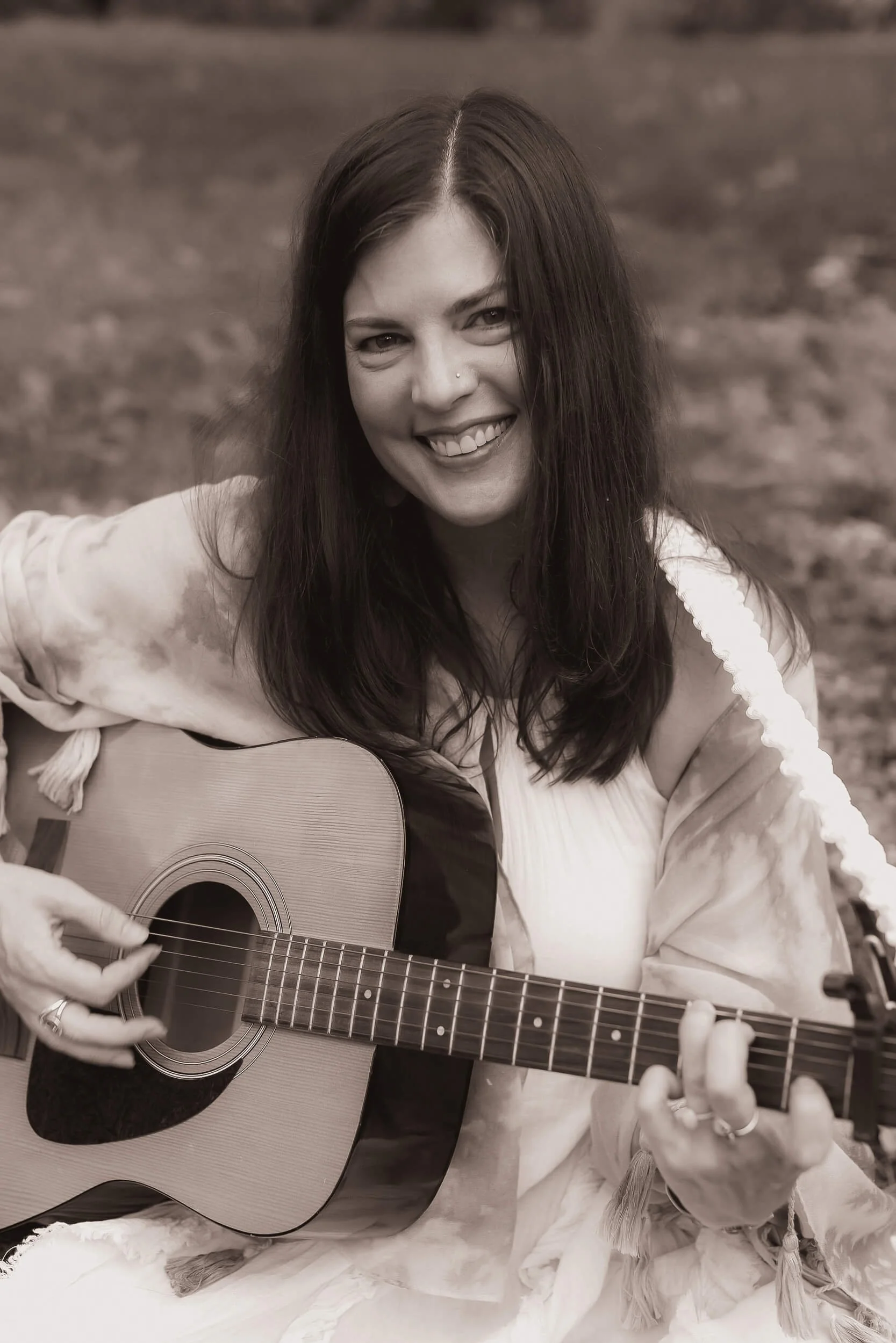 Kristi sitting a field in a dress and playing the guitar