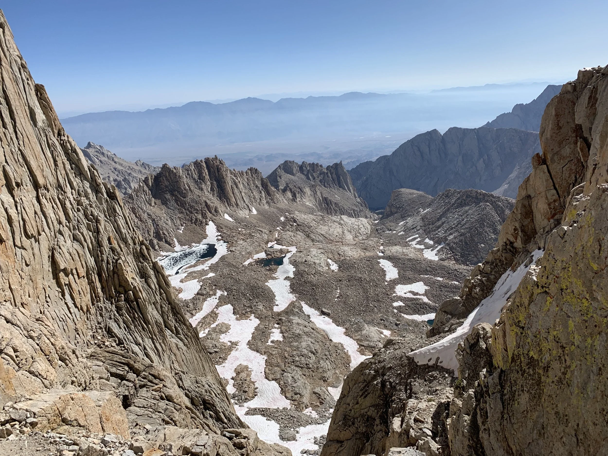 View of rugged mountain terrain with jagged rocks and patches of snow, overlooking a valley with distant mountain ranges and a clear blue sky.