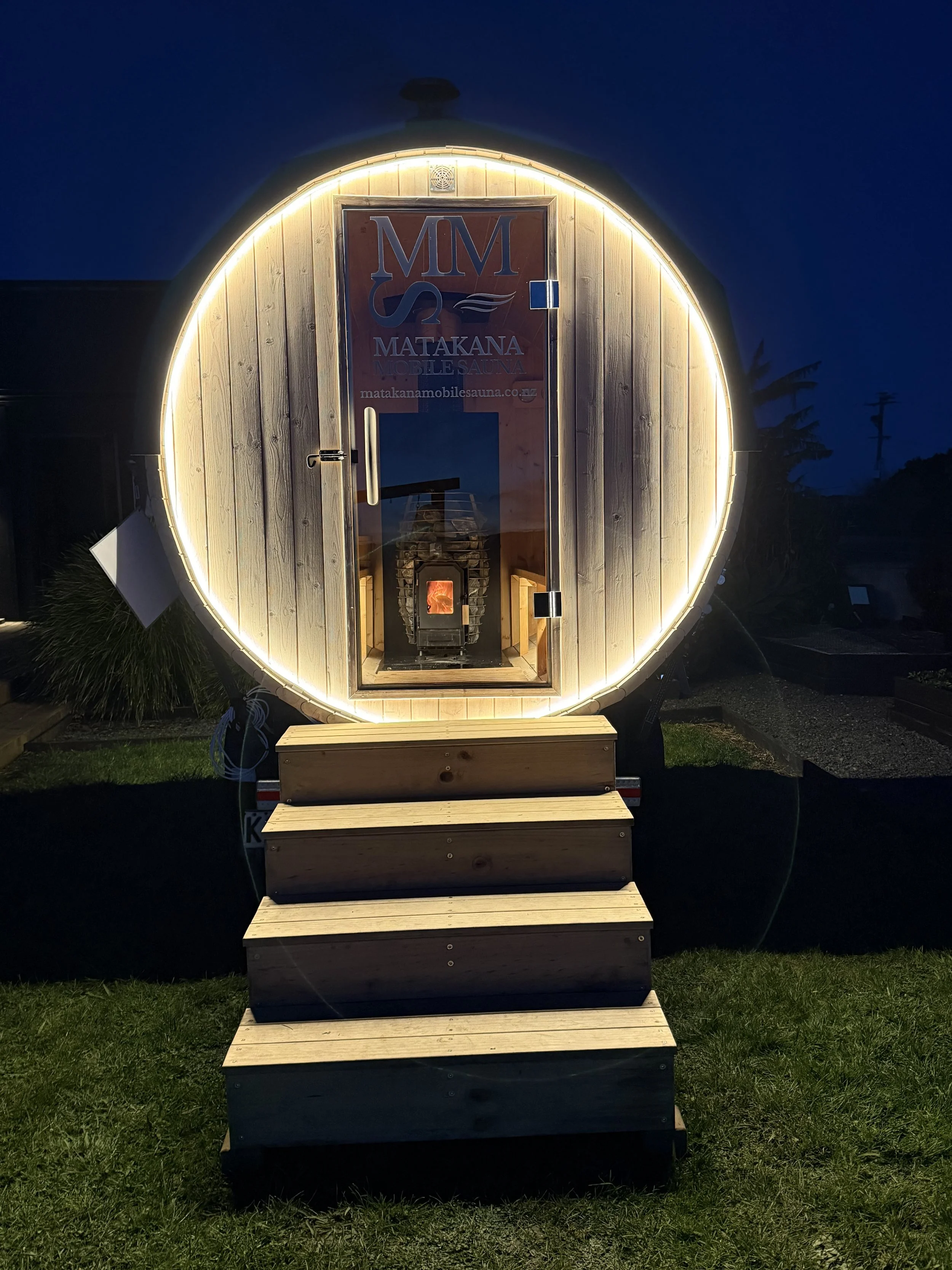 A small, portable sauna with a rounded, wooden exterior and a glowing light border at night. It has a door with a vertical handle, steps leading up to it, and a small wood stove visible inside through the glass door.