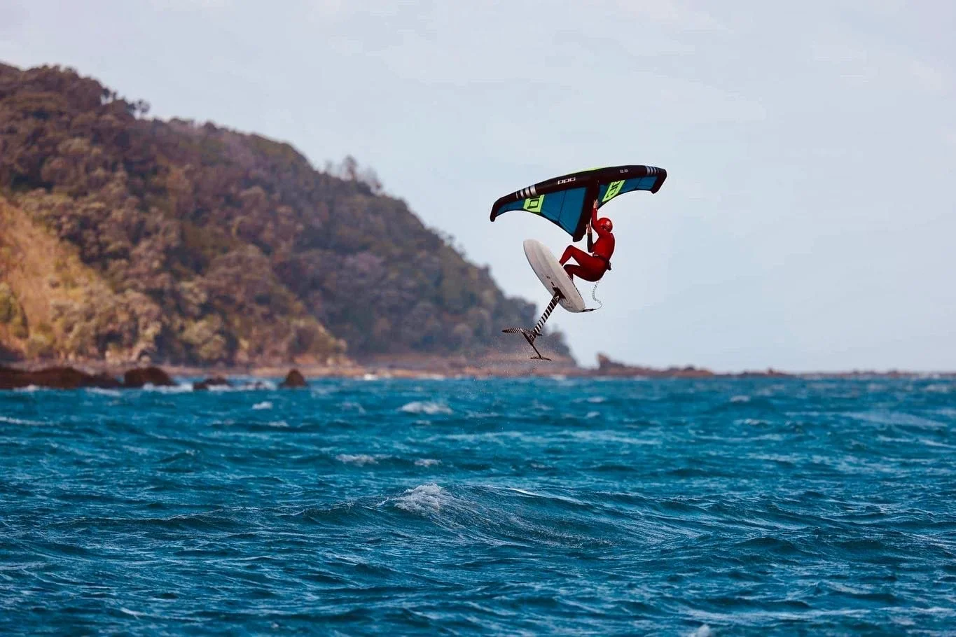A person in a wetsuit is kite surfing over the ocean, performing a jump with the kite and board in mid-air, near a rocky shoreline with green hills.