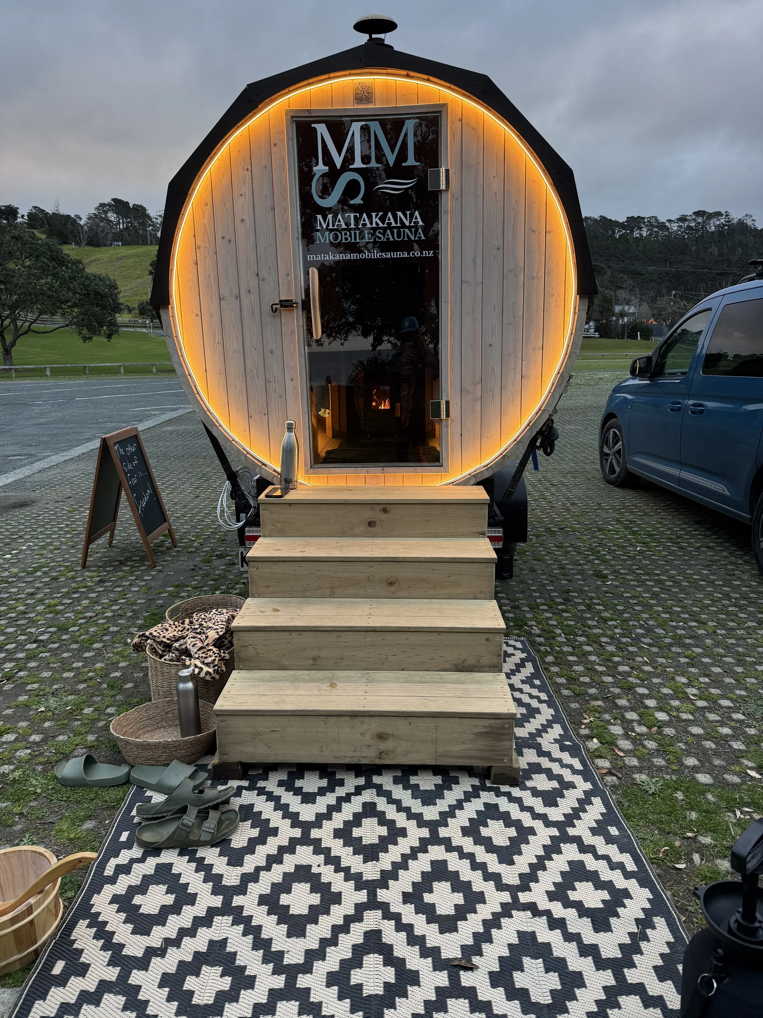 A small, circular sauna with wood paneling and a door with the logo for Matakana Mobile Sauna. It is set on a paved outdoor area with stairs leading up to the entrance. There are sandals, a basket with a water bottle, and a black and white patterned 