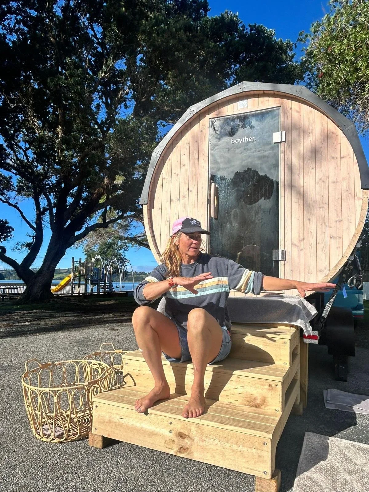 A woman sitting on a wooden bench in front of a small wooden structure with a door, outdoors near a lake with trees and playground equipment in the background. She is barefoot, wearing a baseball cap, a striped long sleeve shirt, and shorts, and is g