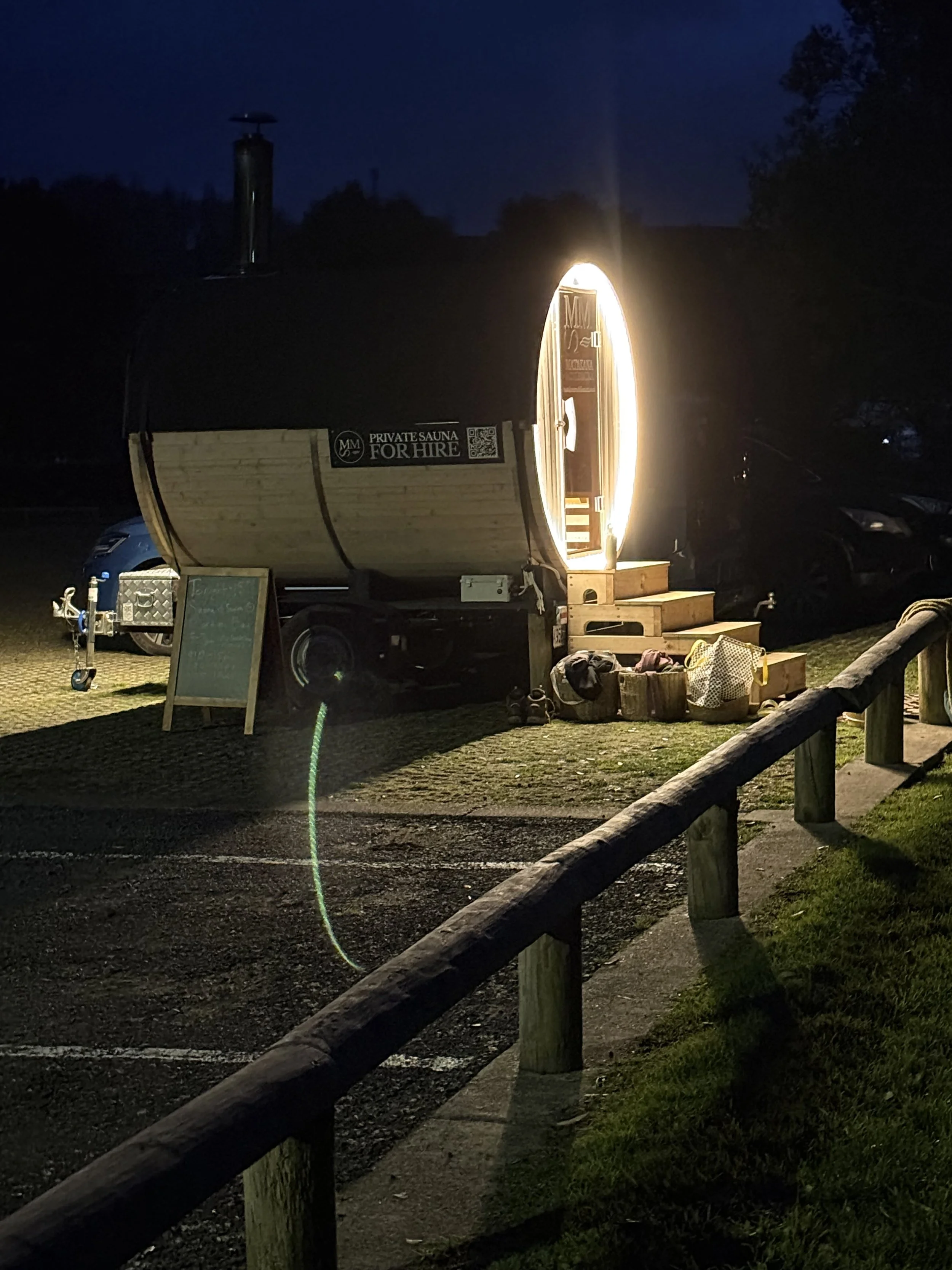 A small wooden sauna trailer with a circular shape is illuminated by a bright light at night, parked on gravel with a chalkboard sign and various bags or items at its base, surrounded by a parking lot with a wooden fence in the foreground and dark sk