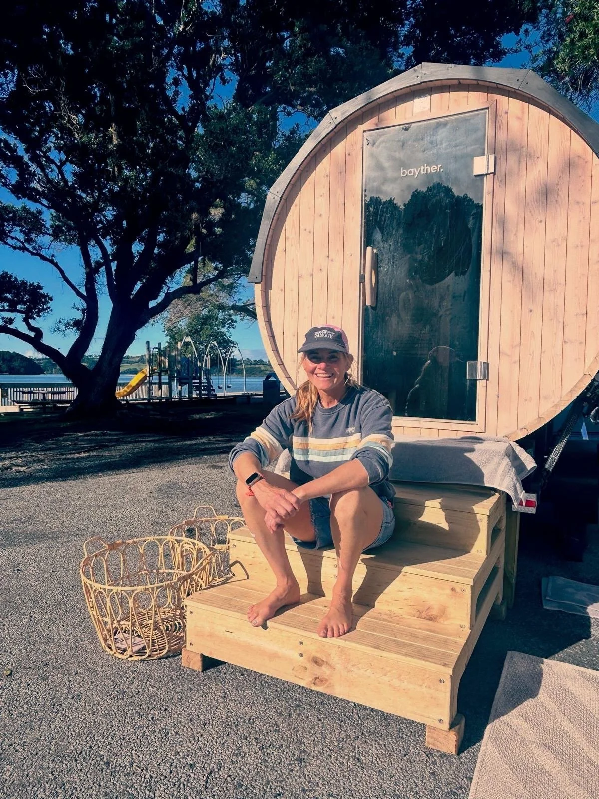 A woman sitting outdoors on a wooden bench in front of a small wooden barrel-shaped structure with a glass door, near a lake with trees and playground equipment in the background. She is smiling, wearing a cap, casual clothes, and has a smartwatch on