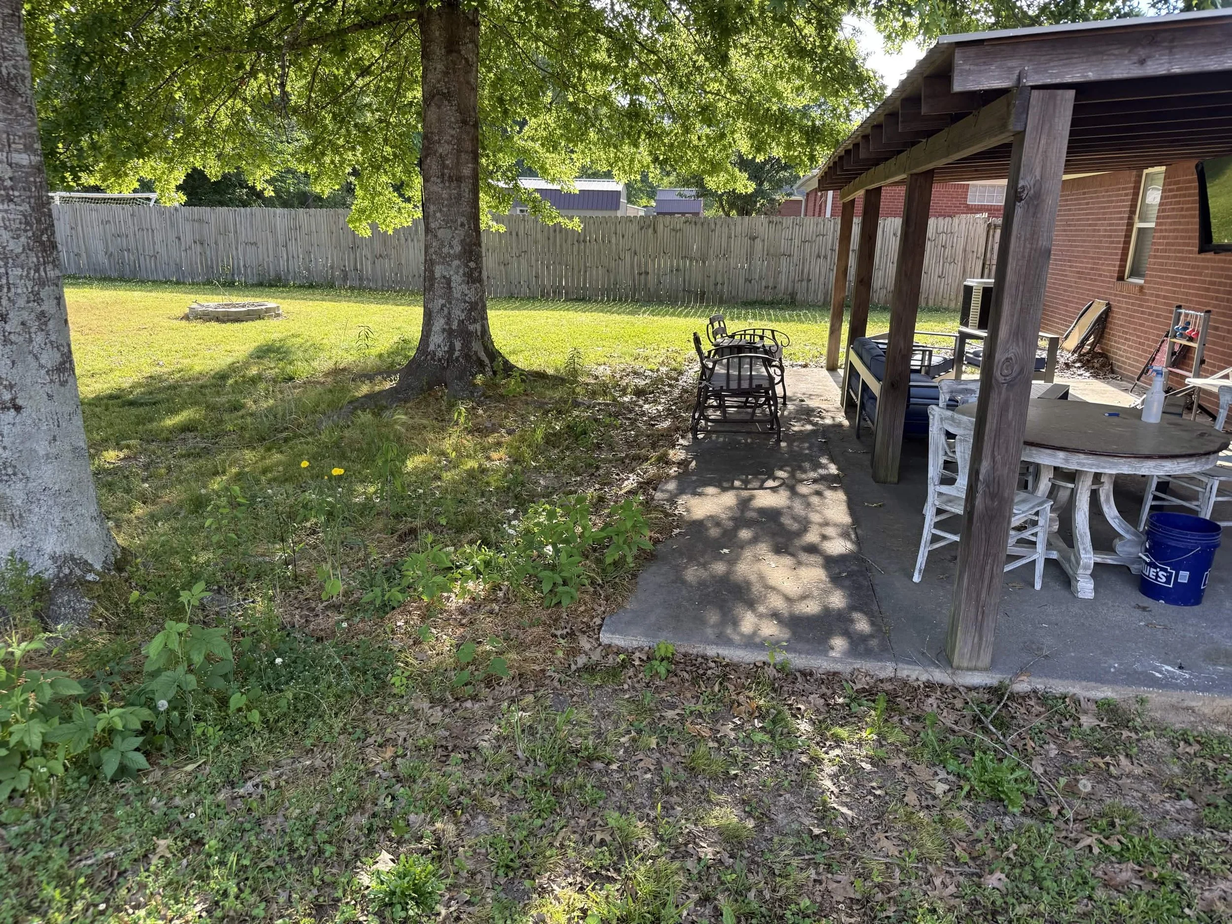 A backyard scene with a large tree, a wooden fence, and a covered patio area with outdoor furniture and a brick house.