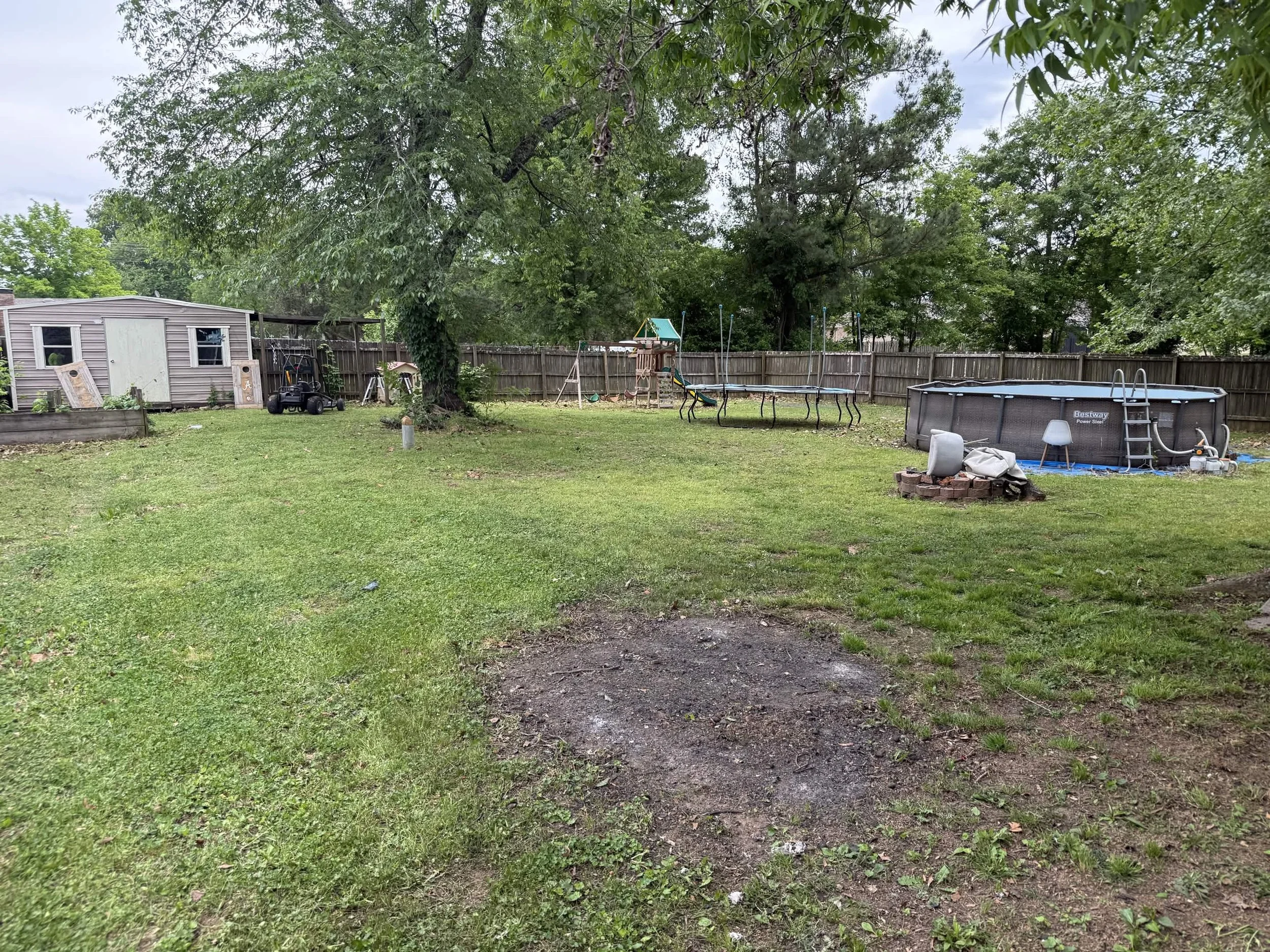 A backyard with a grassy lawn, a wooden fence, a small shed, a trampoline, and various playground equipment surrounded by trees.